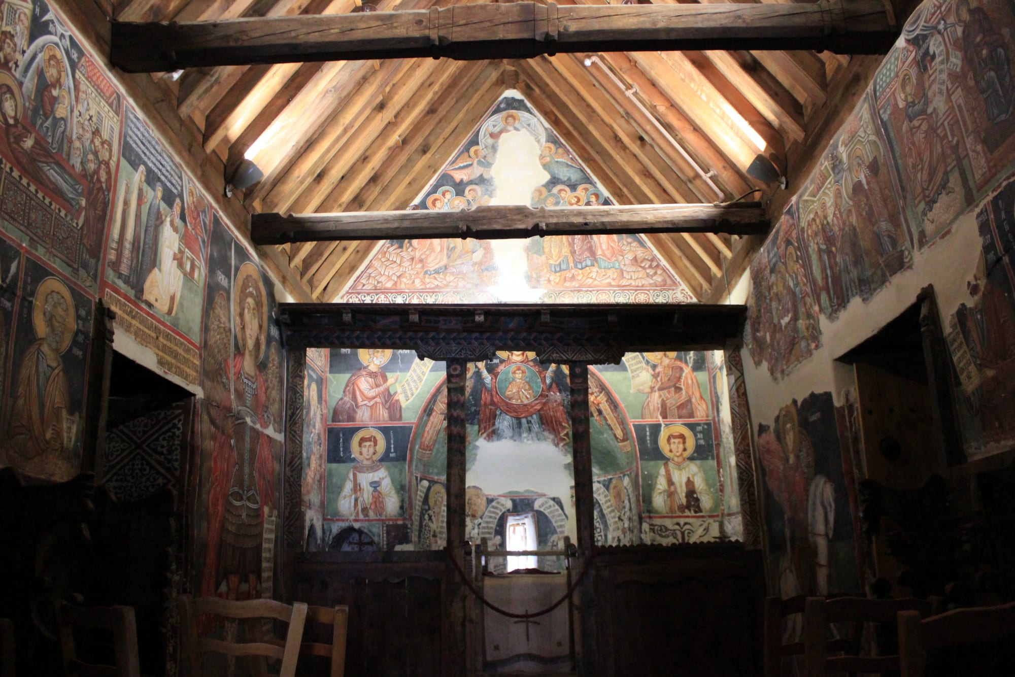 The interior of a small church with a pitched timber roof, its walls and wooden screen covered in brightly coloured paintings of saints with golden haloes