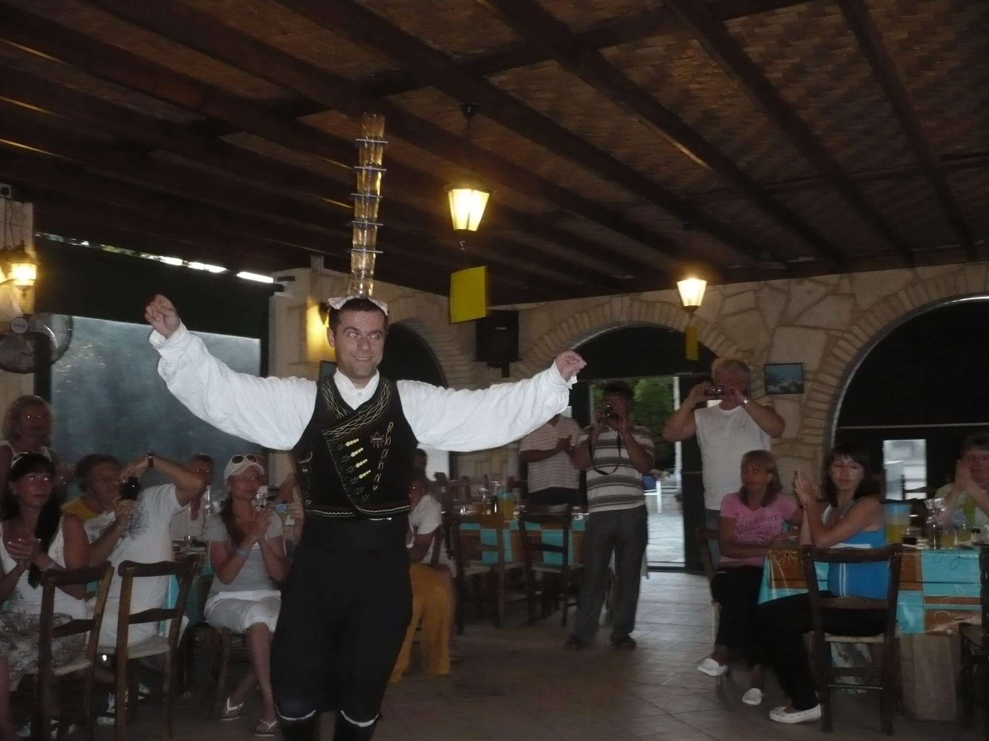 A man in a white shirt, black embroidered waistcoat and black breeches dances with his arms outstretched and a tall tower of stacked glasses balanced on his head, while seated diners watch and take photographs