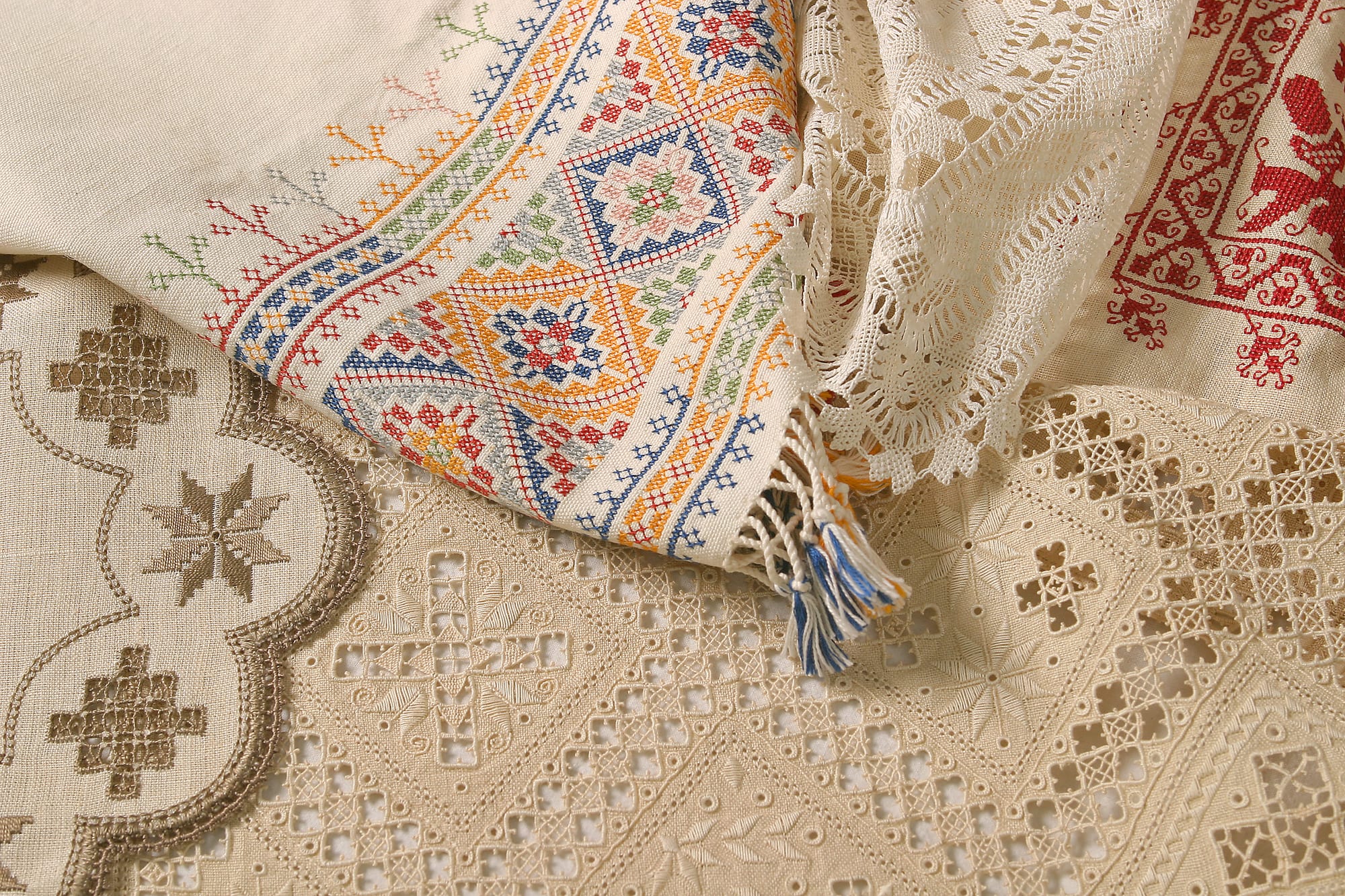 Folded pieces of cream linen needlework laid over one another, some with open geometric cut-work and lace edgings, one with bright cross-stitch borders in blue, orange and red, and one with red embroidered motifs