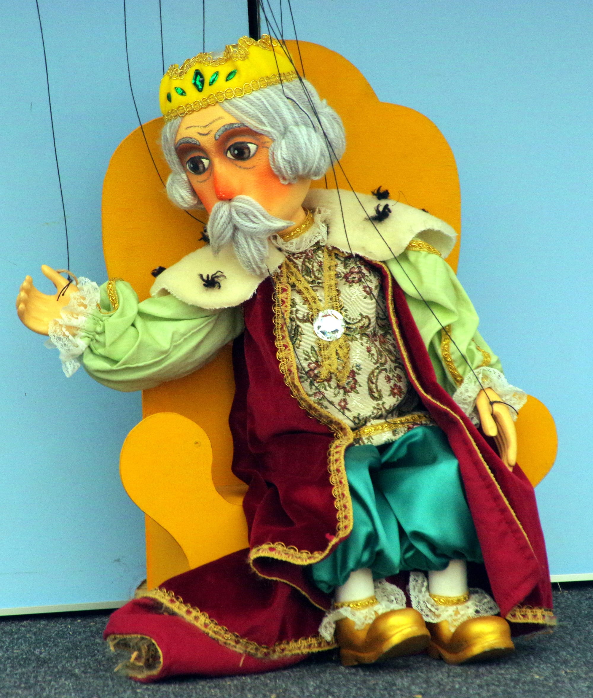 A wooden marionette of an elderly king with a gold crown, grey hair and moustache, a red velvet cloak, green sleeves and gold shoes, seated on a yellow wooden throne with strings rising from his head and hands
