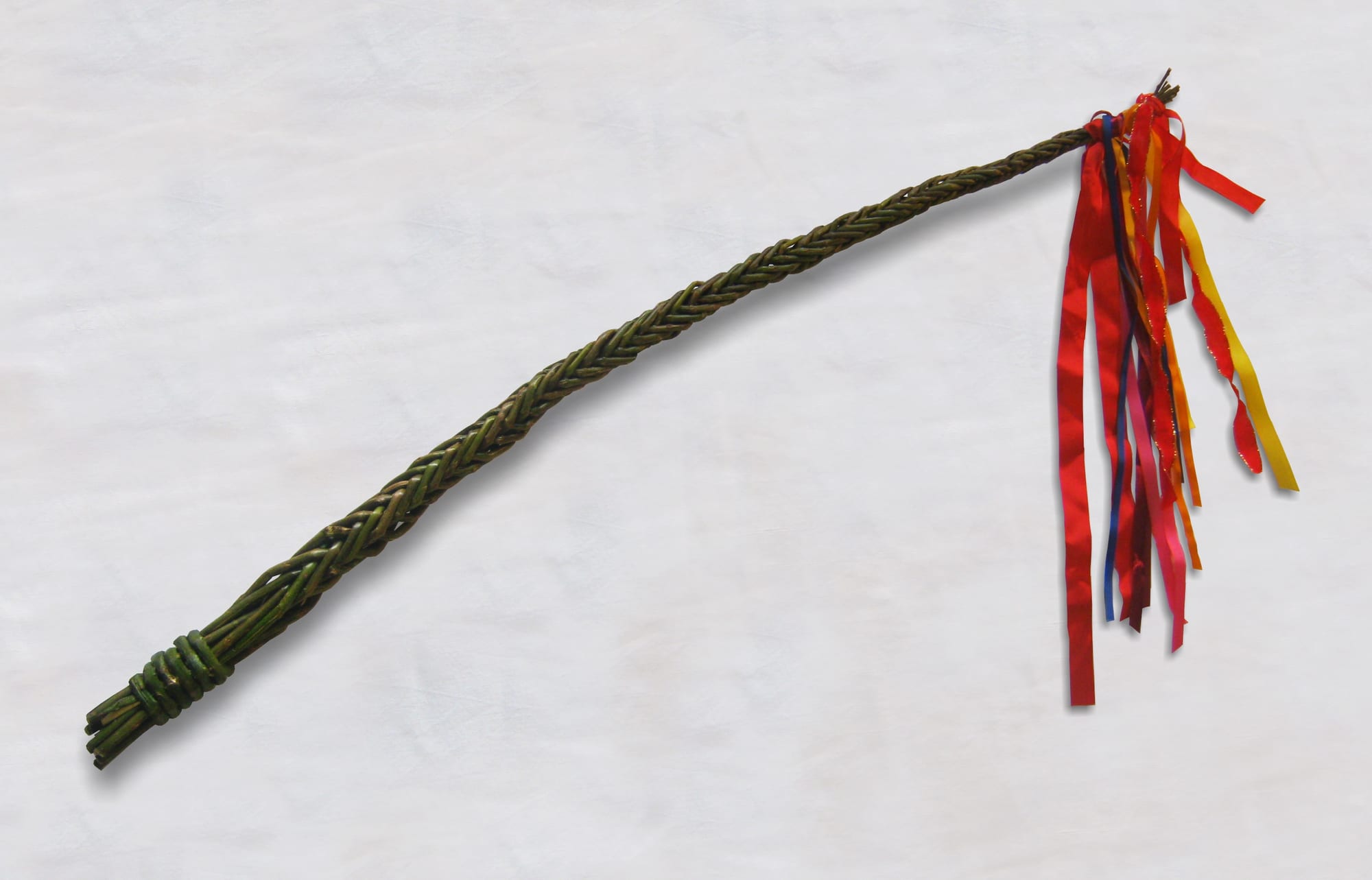 A long whip of green willow rods braided together, with a bunch of red, orange, yellow, pink and blue ribbons tied at the tip, lying on a white background