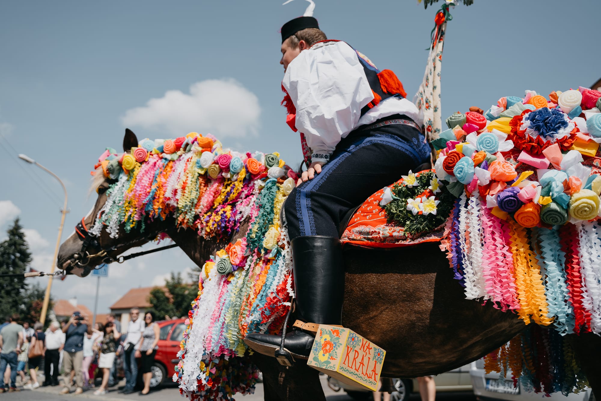 A young rider in a white shirt, black cap and embroidered blue breeches sits on a brown horse whose mane and saddle are covered in hundreds of colourful handmade paper roses and ribbons, with spectators watching from the roadside