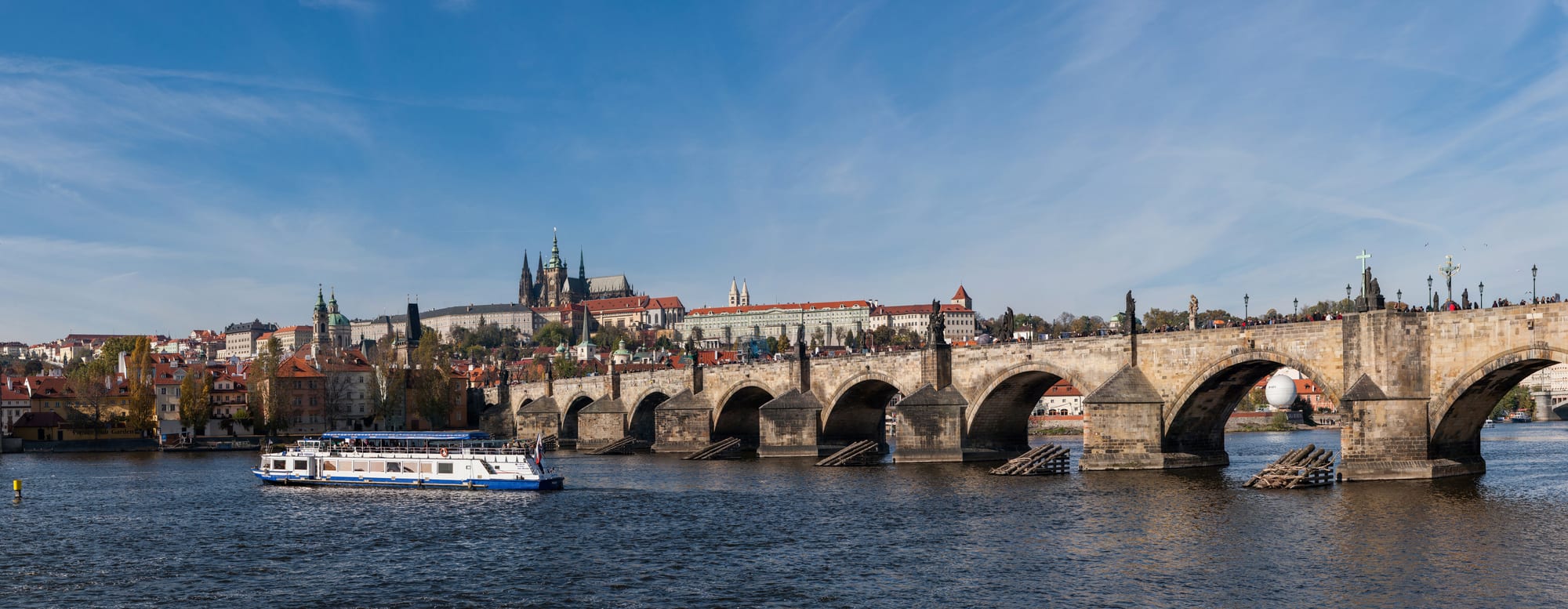 A medieval stone bridge with statues leading to a hilltop castle crowned by a Gothic cathedral spire
