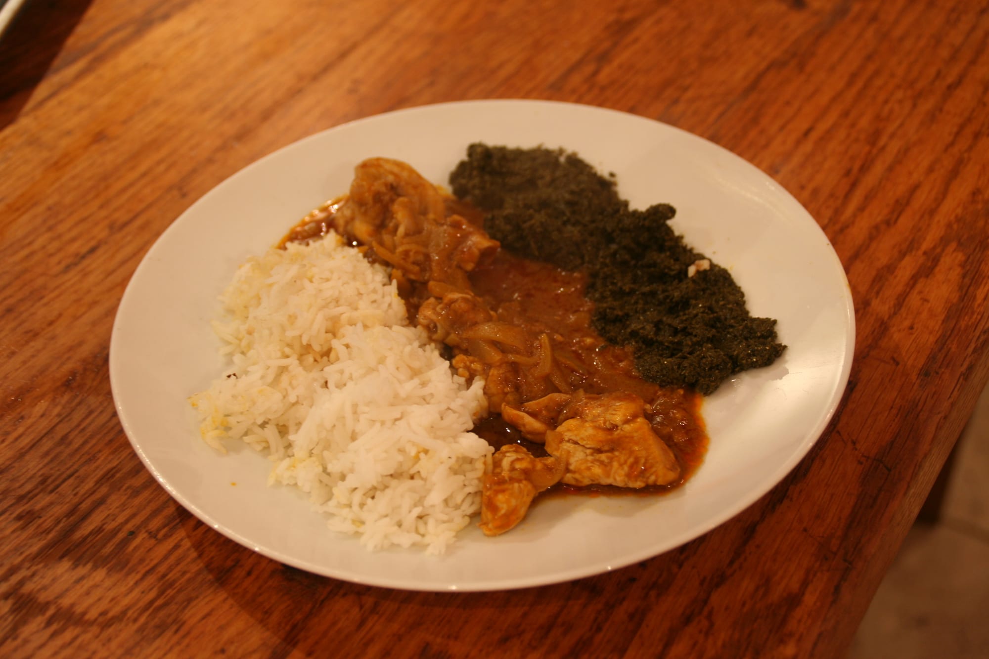 A plate of chicken in an orange palm-nut sauce, served with rice and dark green cassava leaves