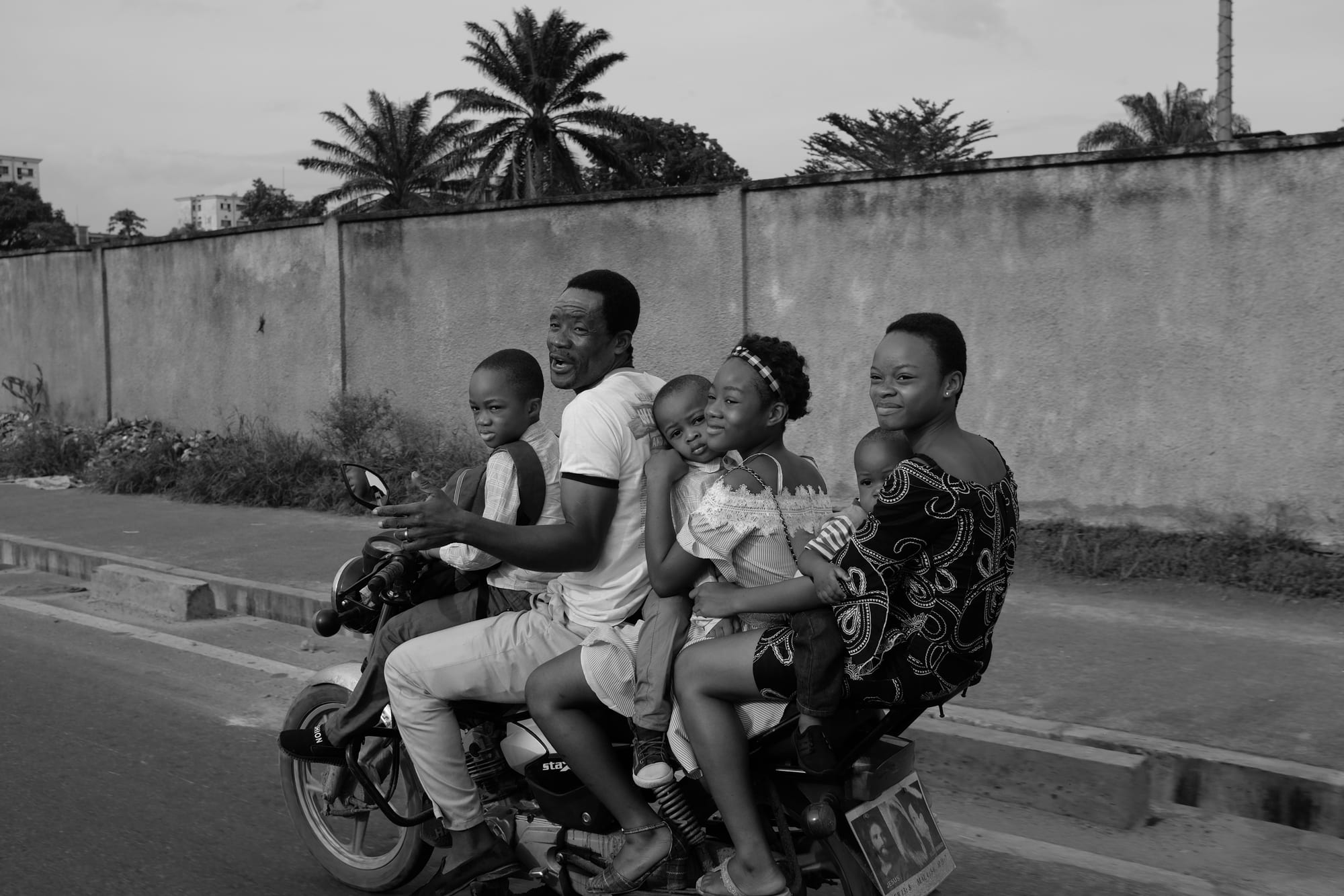 A family of six, a father driving, riding together on one motorbike past a long roadside wall lined with palm trees