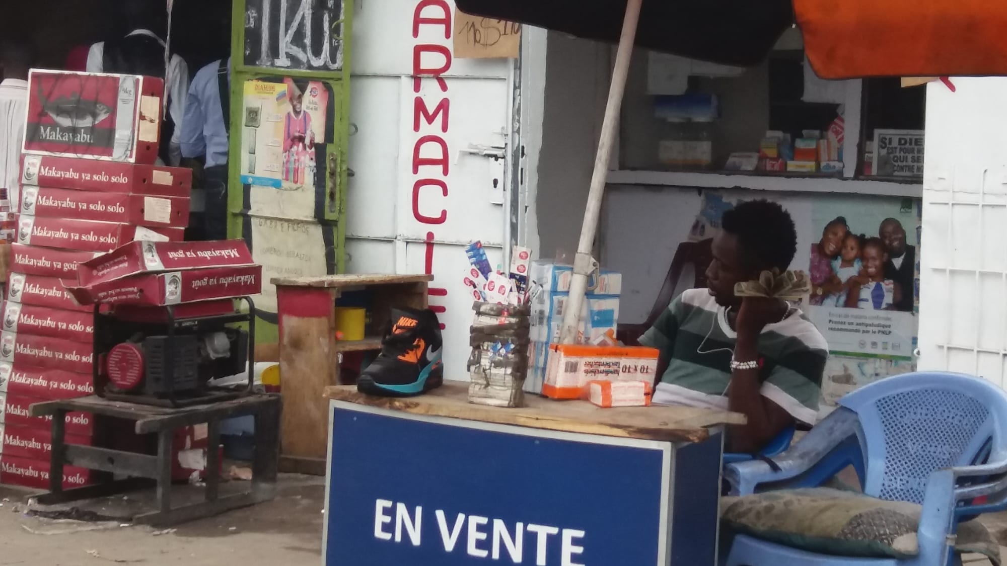 A roadside vendor's stall in Kinshasa piled with shoes, phone cards and boxed goods beside a pharmacy shopfront, with the seller seated nearby