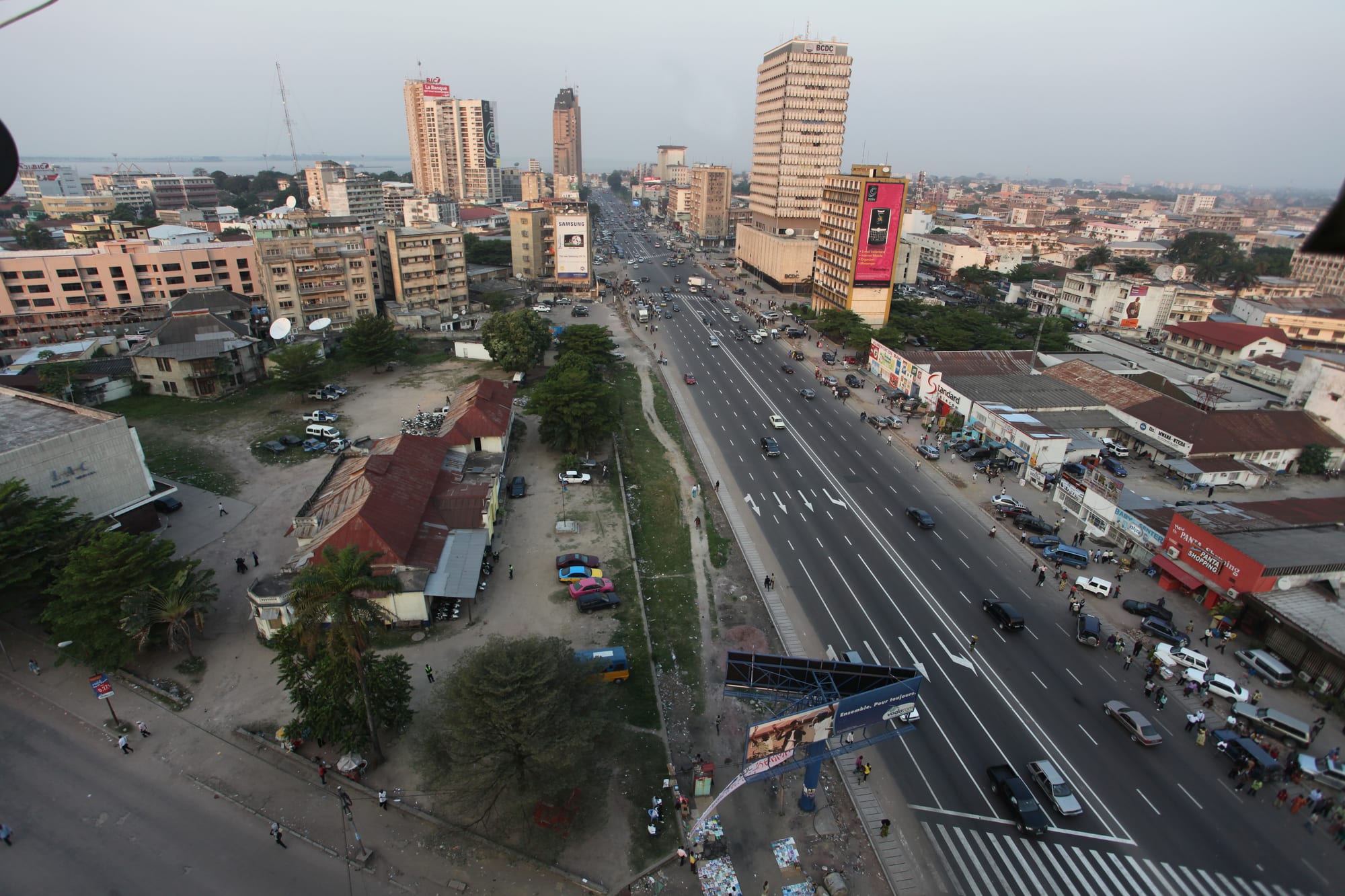 An aerial view of a wide multi-lane boulevard in Kinshasa lined with tower blocks, cars and roadside stalls