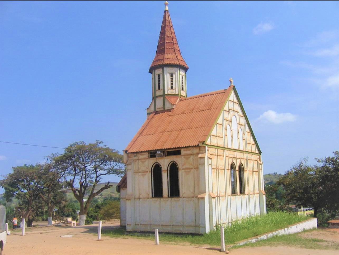A small cream-coloured church with a steep red spire, a rust-red corrugated roof and tall arched windows, standing on a bare earth square under a blue sky