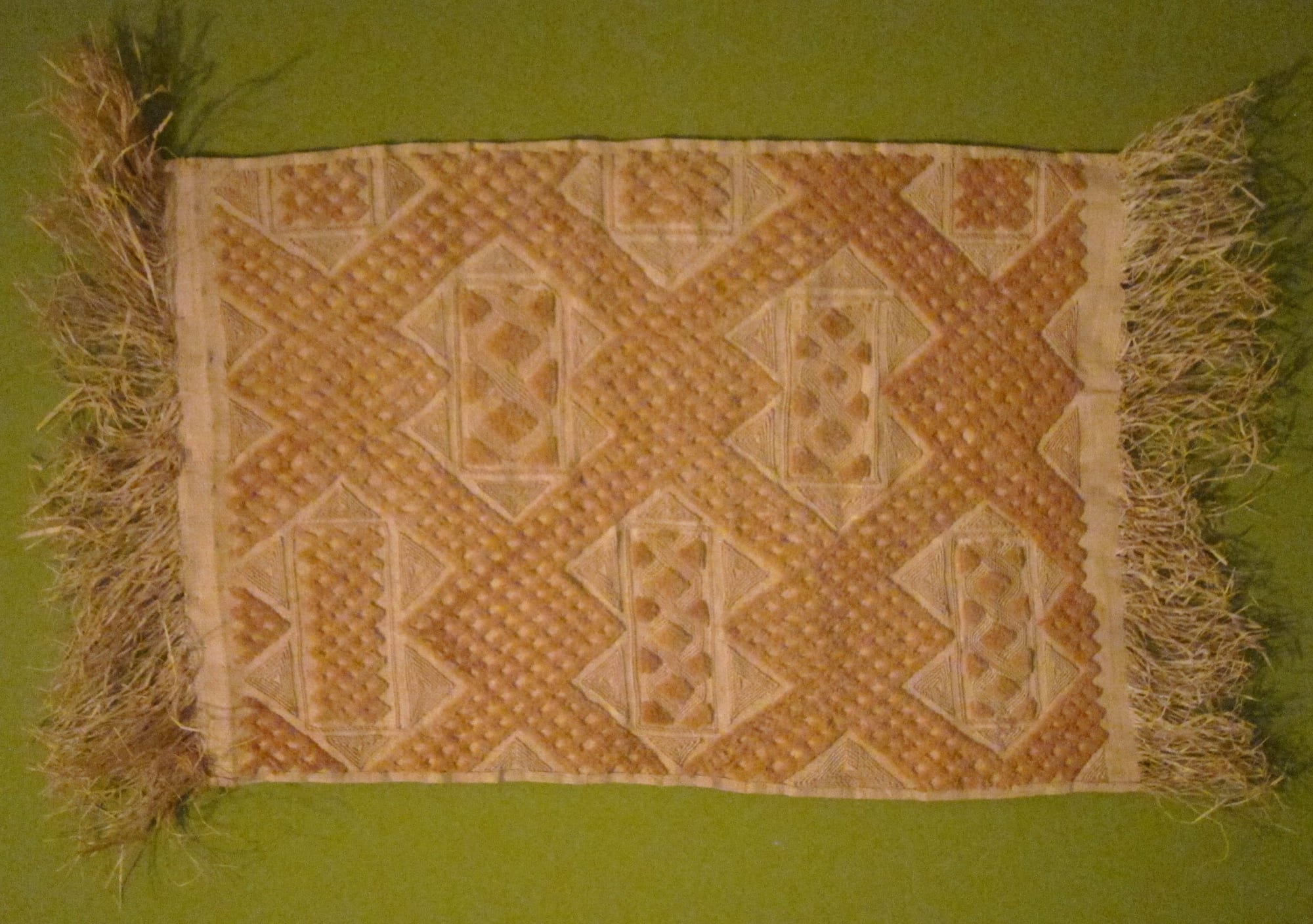 A rectangular panel of pale raffia cloth with fringed ends, covered in raised embroidered geometric patterns of diamonds, triangles and interlocking bands in golden and cream tones