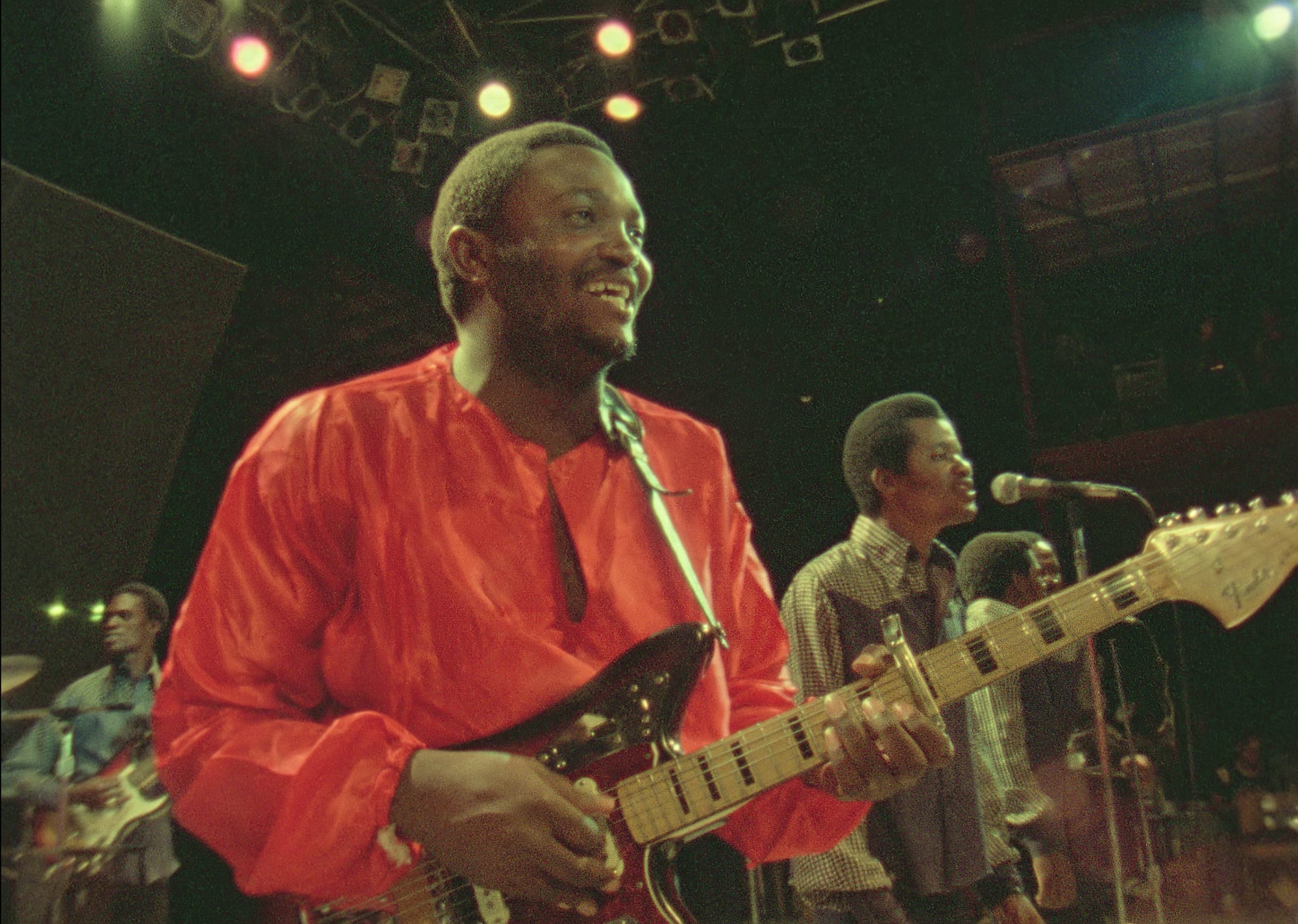 A smiling guitarist in a loose orange shirt plays an electric guitar on a stage under coloured lights, with a singer at a microphone and another guitarist behind him