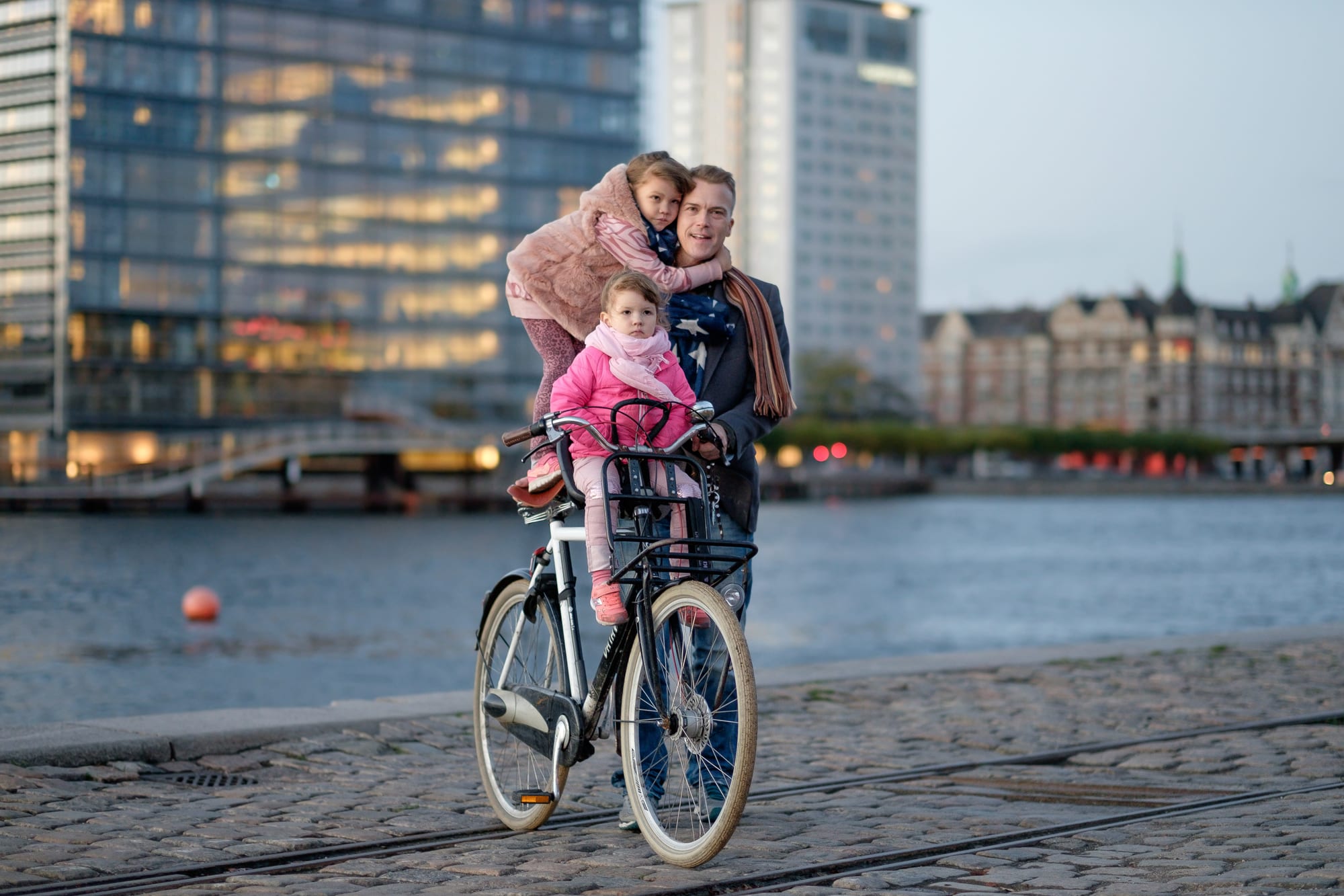 A father cycling with one daughter riding on the back rack and another in a front-mounted child seat, a waterfront and office towers behind them