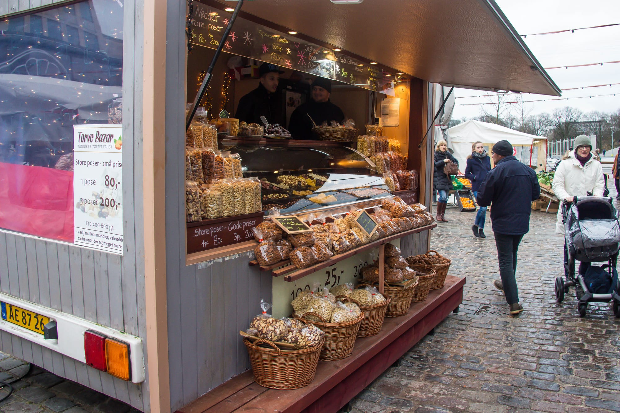 A market stall stacked with bagged nuts and dried fruit, a vendor serving customers, with shoppers and a pram passing on wet cobblestones