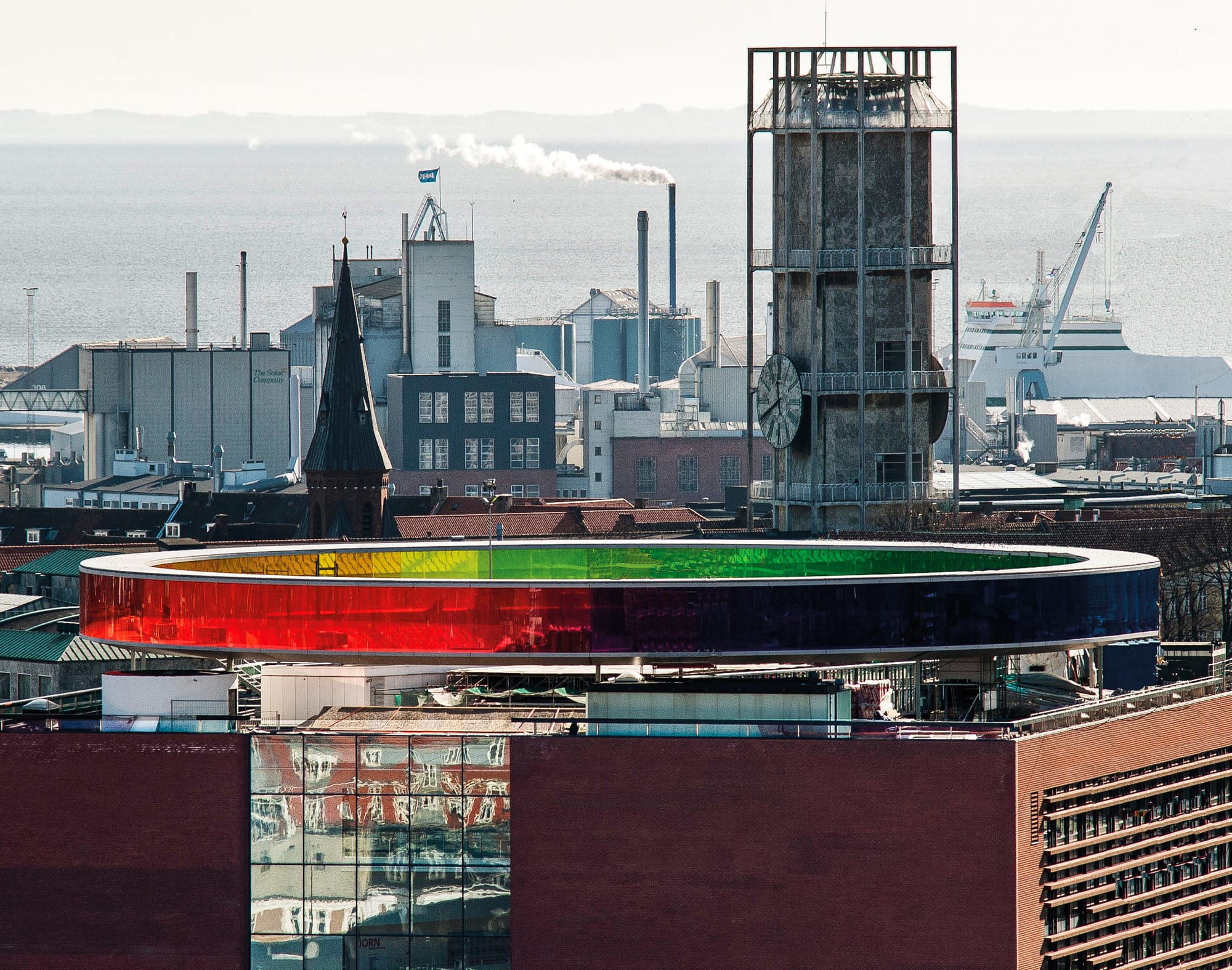 A circular walkway of rainbow-coloured glass on the roof of a museum, city beyond