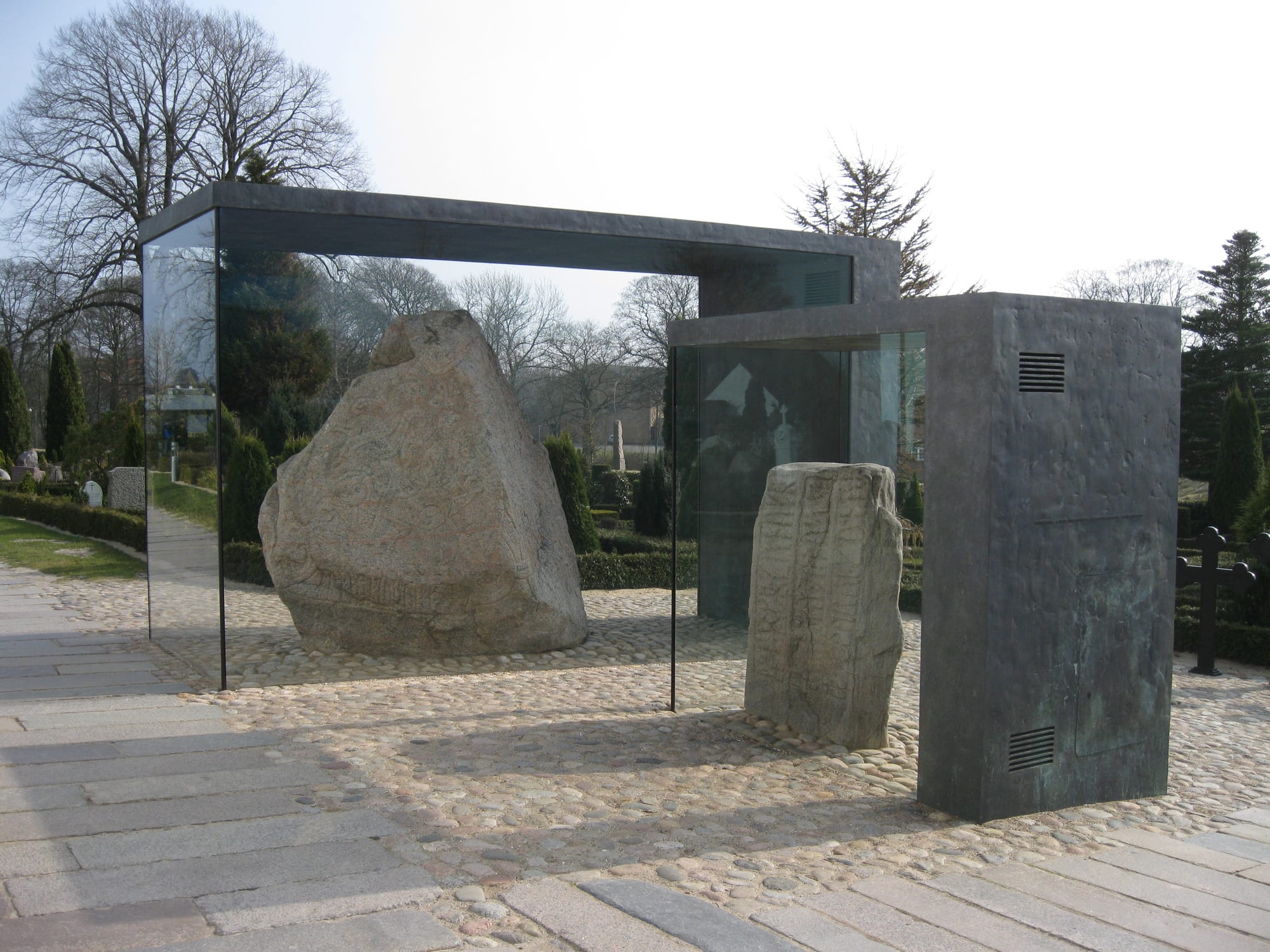 A large carved granite runestone under a glass case beside a whitewashed church and burial mounds
