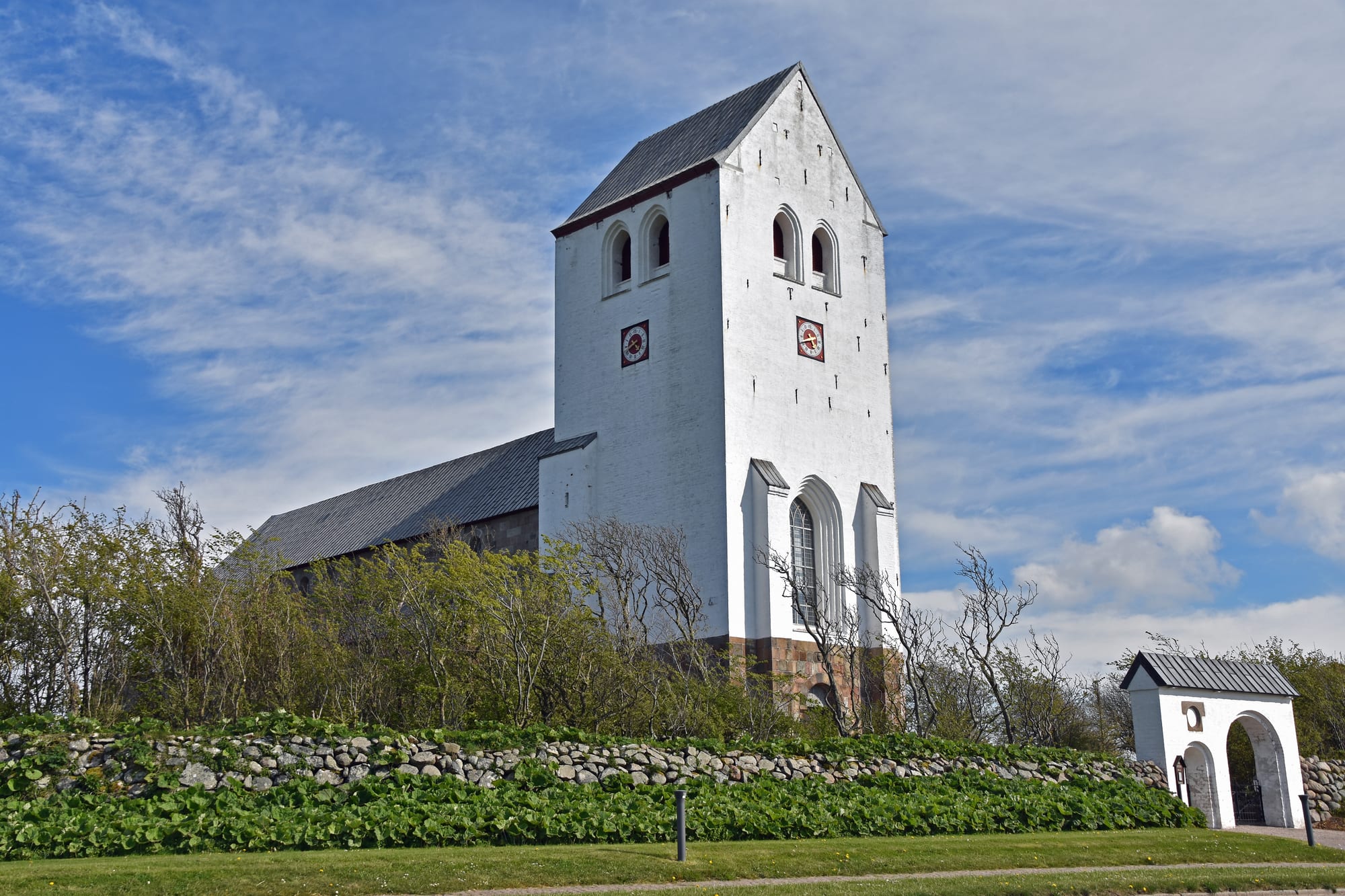 A large whitewashed church tower with a stepped gable and small arched windows rising above a stone-walled churchyard, with a white gateway to one side and a blue sky with thin cloud