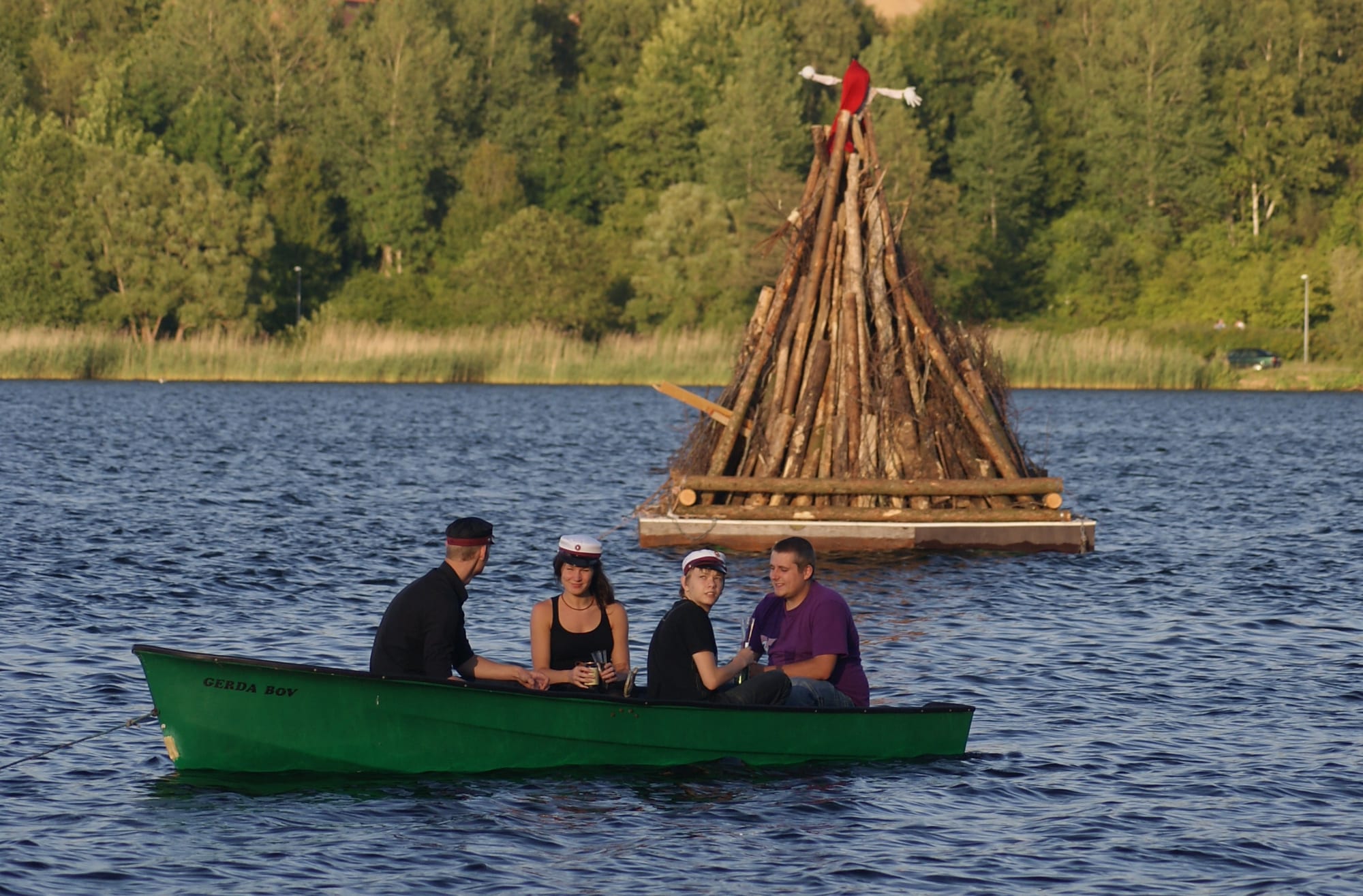Four young people, three wearing white-topped student caps, sit in a green rowing boat on a lake beside a tall stack of logs on a floating platform, topped by a figure made of old clothes