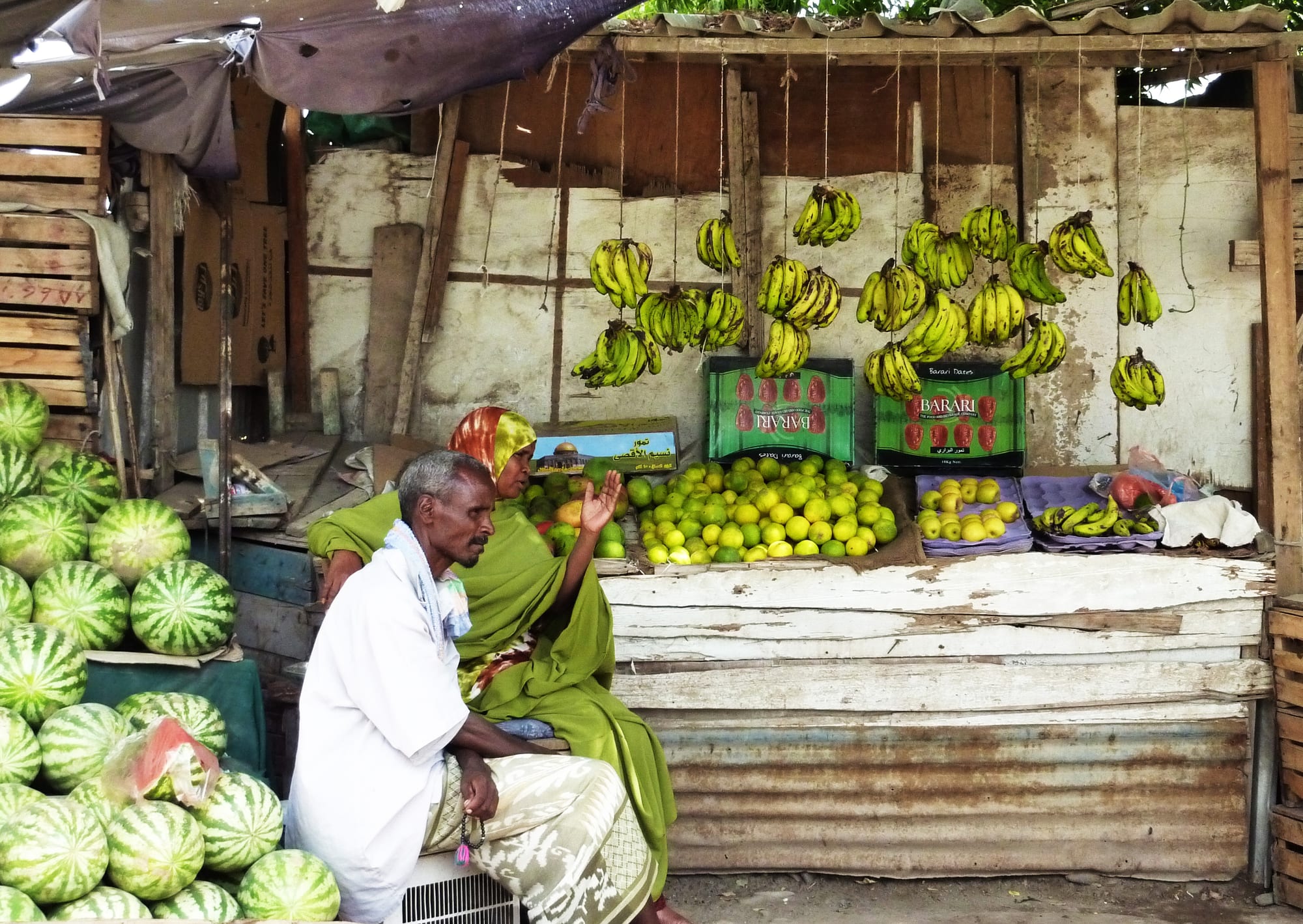 A market vendor sitting beside a fruit stall with watermelons, limes and bananas hanging from the roof of a wooden stall