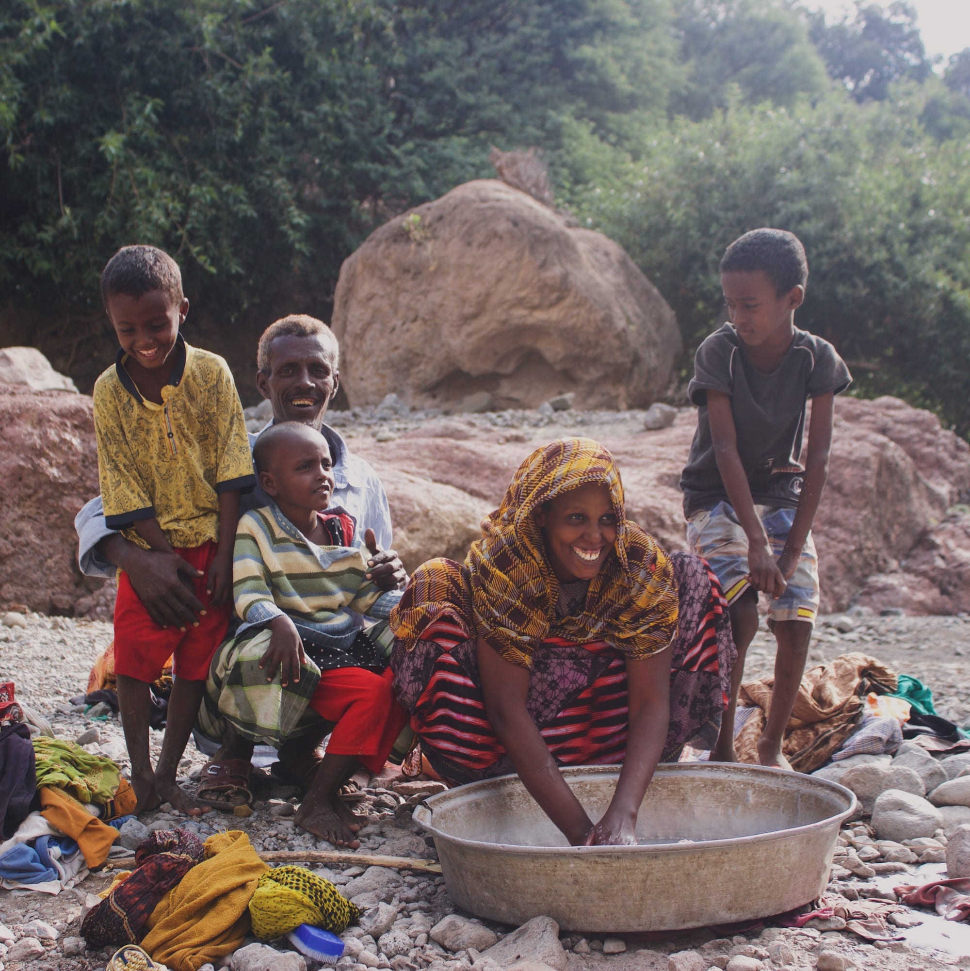 A family group, including a woman washing clothes in a large metal basin and several children and a man watching, on a rocky dry riverbed