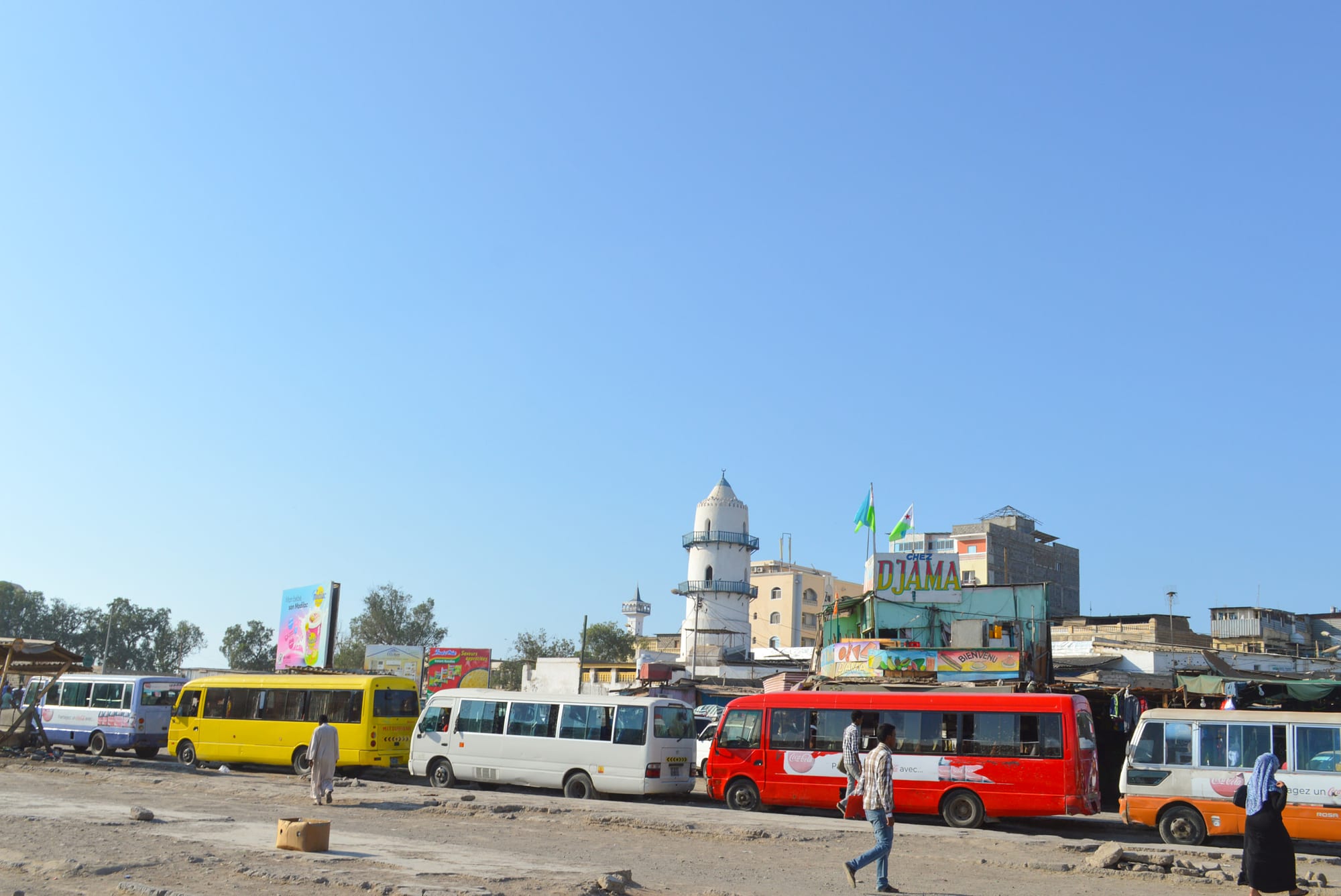 A row of colourful minibuses lined up at a bus station in Djibouti City, with pedestrians crossing and a mosque minaret in the background