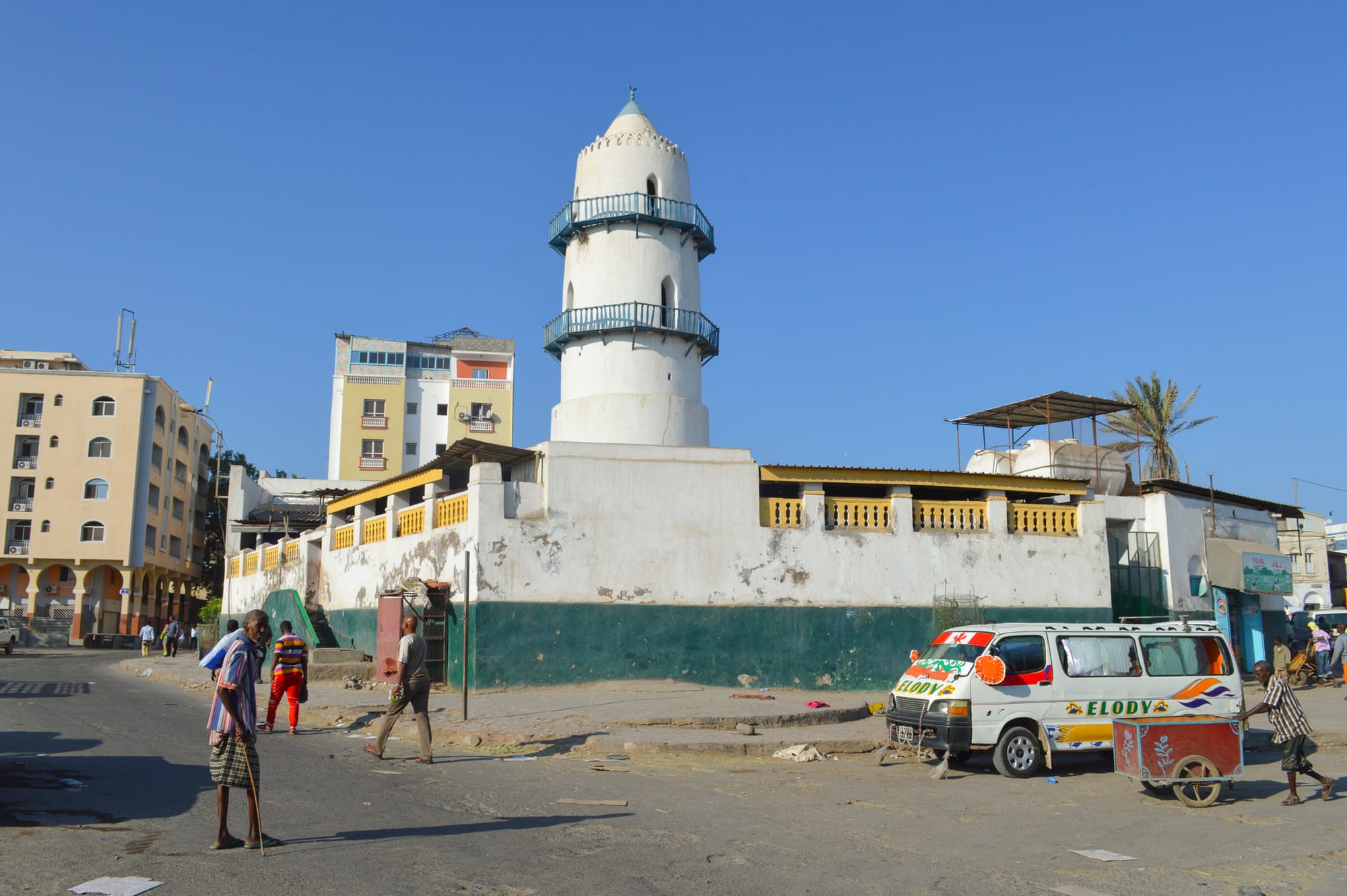 A white mosque with a slender minaret rising above a busy market street of low buildings