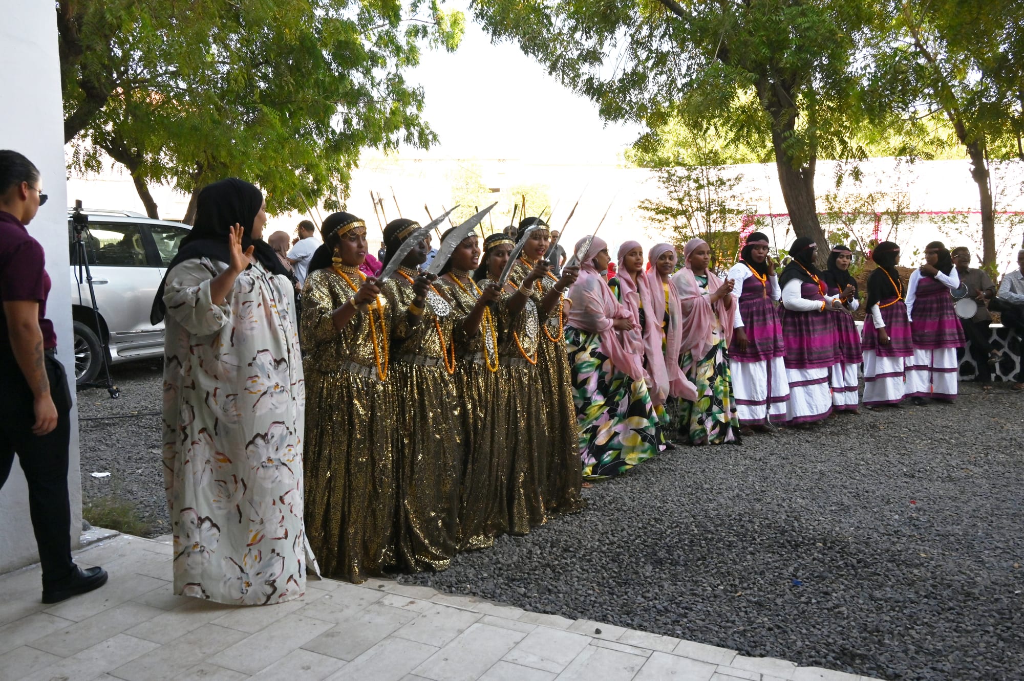 A line of women in gold, patterned and purple traditional dresses with headscarves and amber necklaces, several holding up curved blades, performing in a courtyard under trees