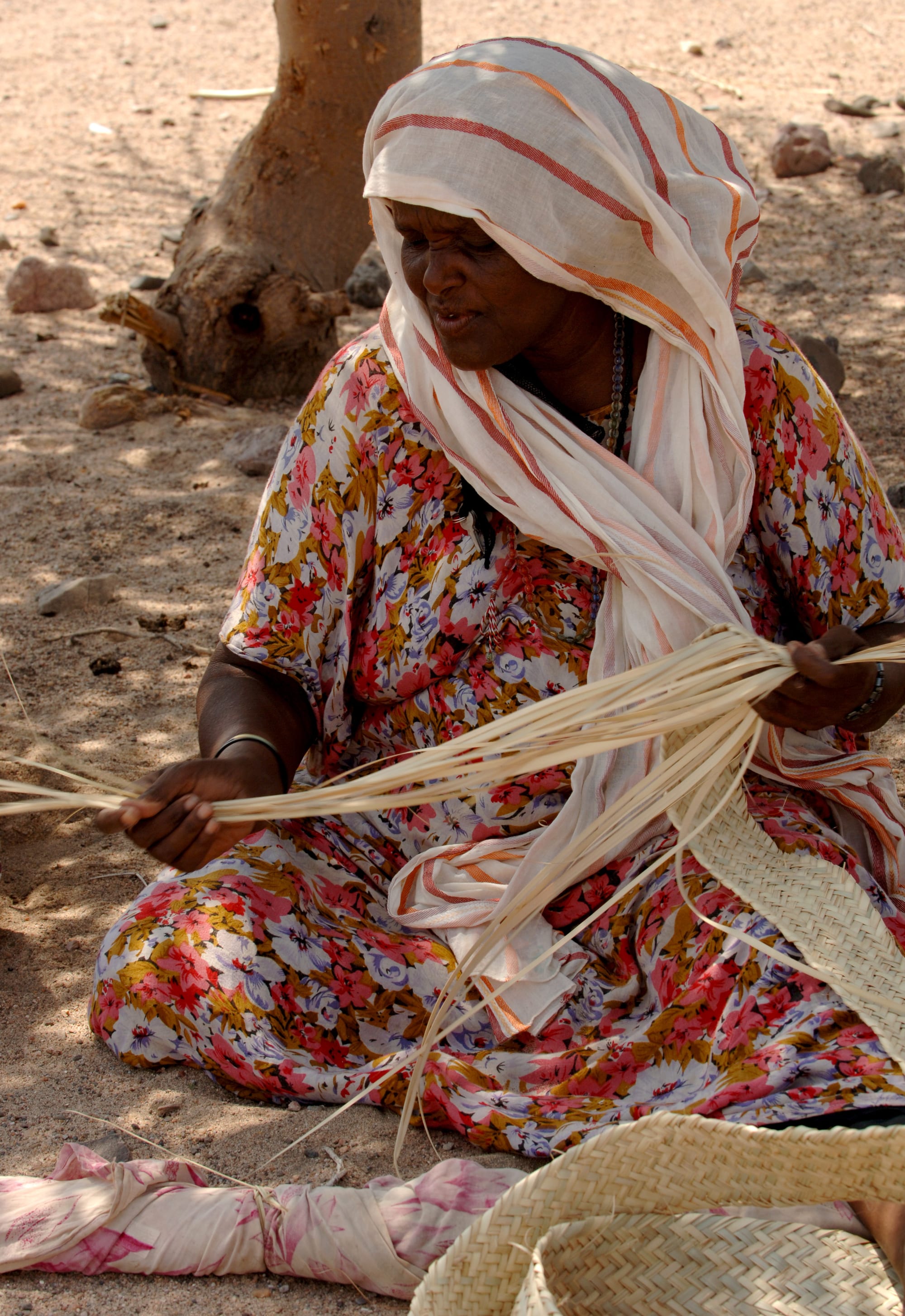 A woman in a floral dress and a striped headscarf sits on sandy ground in the shade of a tree, weaving long pale fibre strips into a basket