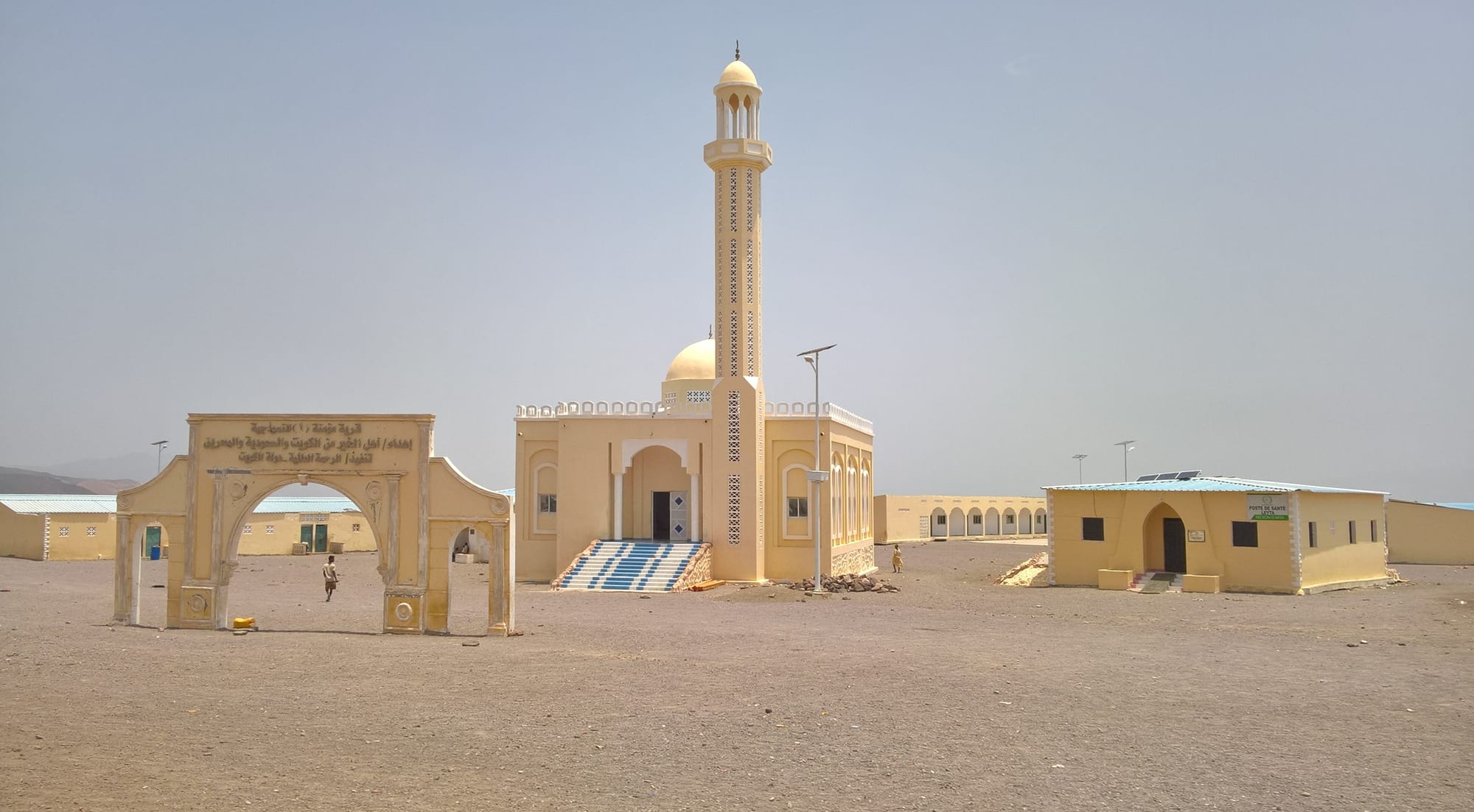 A cream-coloured mosque with a dome and a tall slender minaret stands on open sandy ground beside an arched gateway and low buildings, under a hazy sky