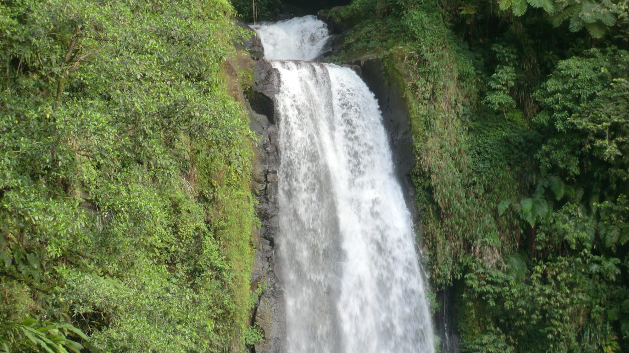 Two waterfalls plunging side by side down a black cliff into a jungle gorge strewn with boulders