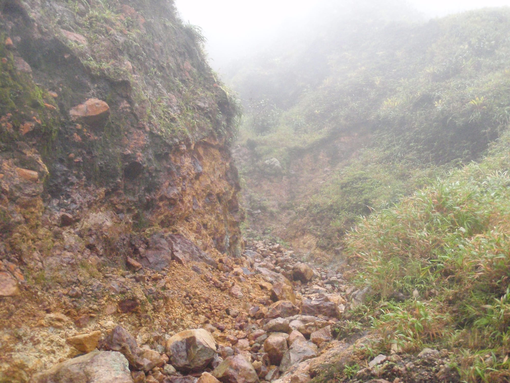 A hiker descending toward a barren valley of grey and yellow sulphur-stained rock with steam rising, green ridges around