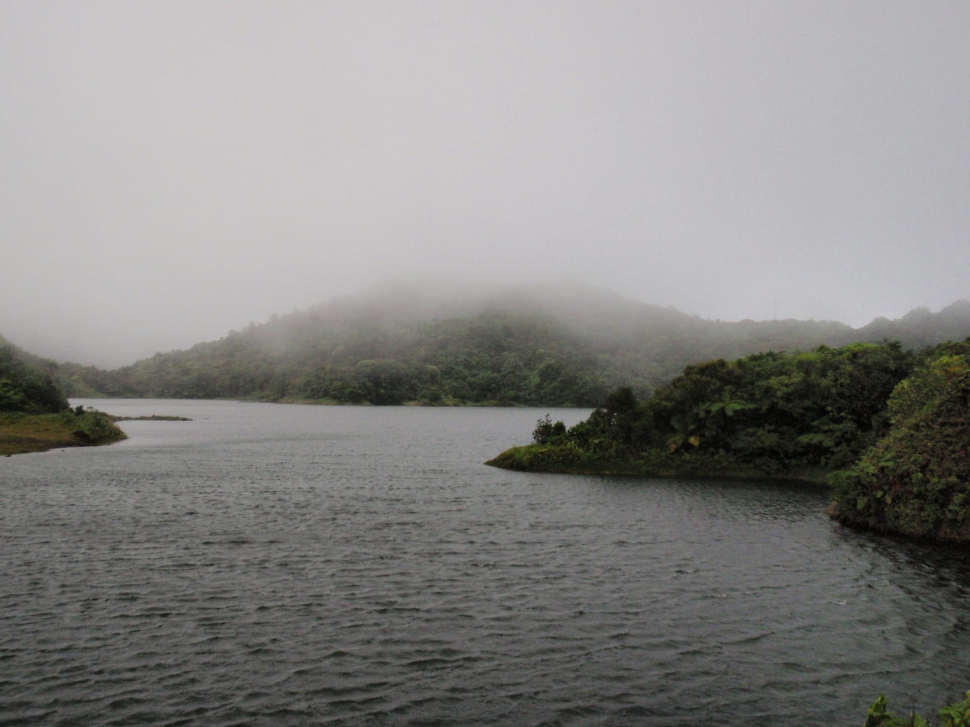 A calm crater lake surrounded by steep green ridges under a grey sky