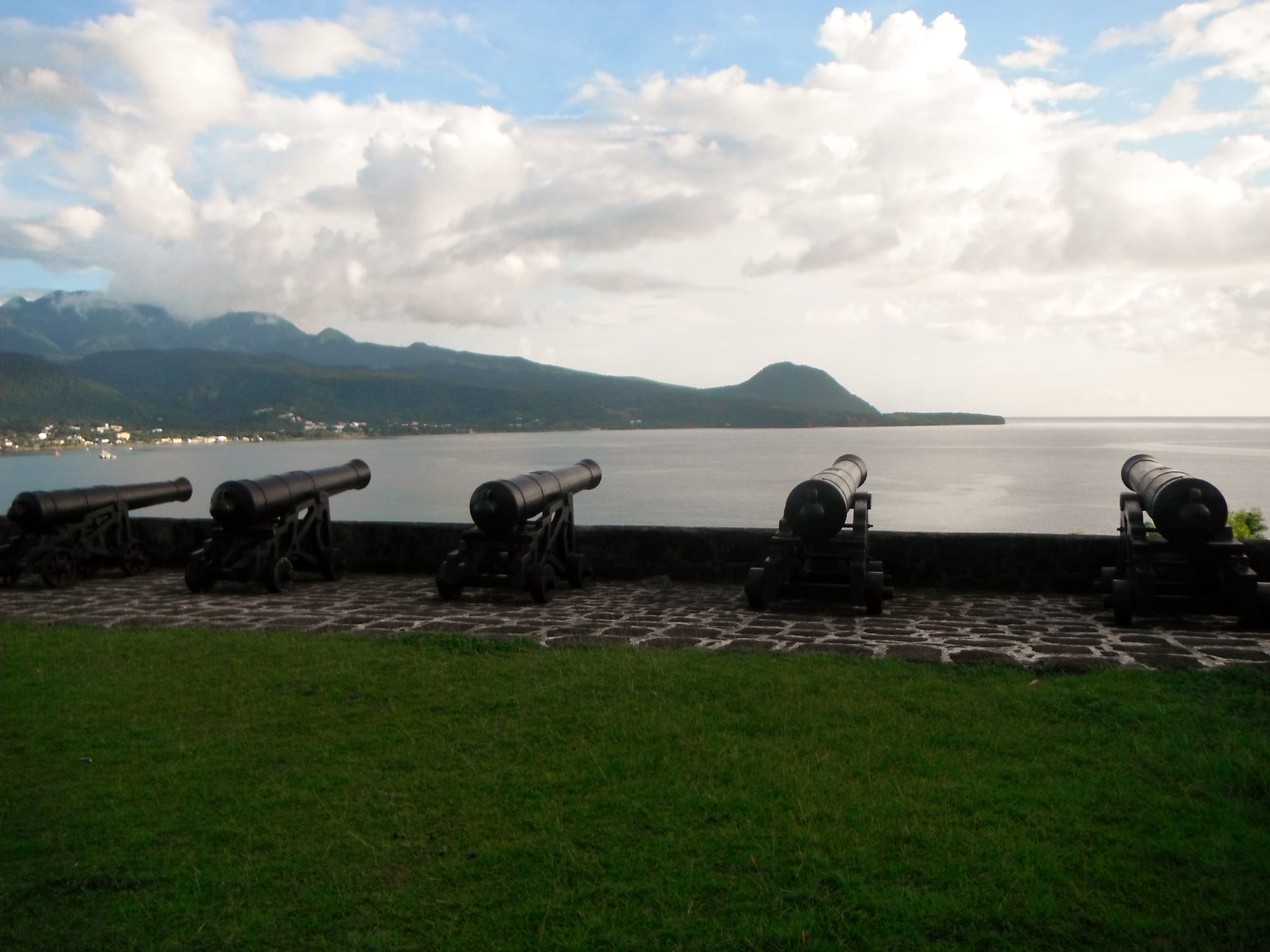 A row of black cannons on low wheeled carriages lined up along a low stone parapet above a calm bay, with green mountains and clouds beyond