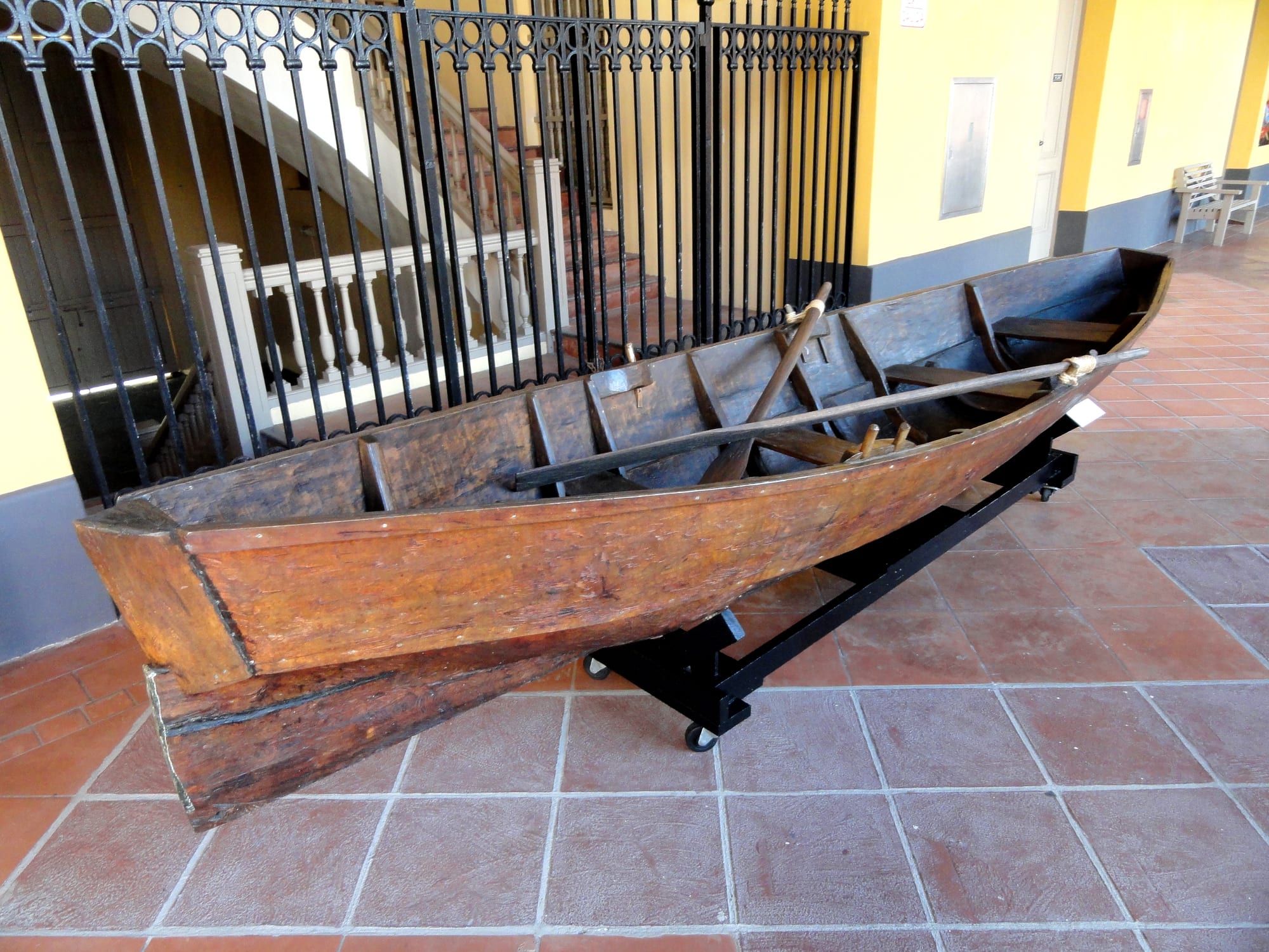 A long wooden canoe hollowed from a single tree trunk, with plank seats and a paddle laid across it, displayed on a stand in a museum arcade