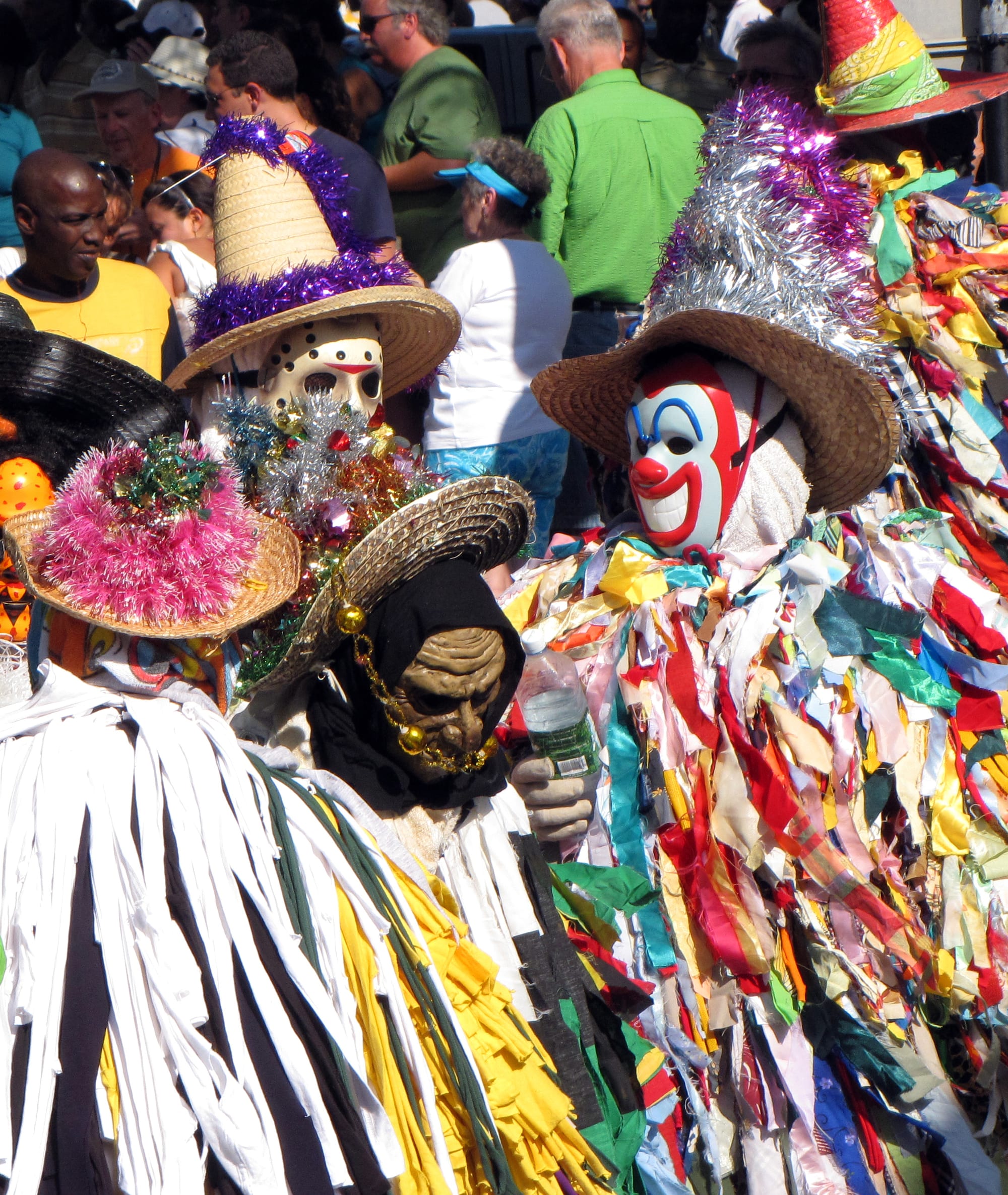 Masqueraders in costumes of layered, multicoloured cloth strips, with masks and tinsel-trimmed straw hats, moving through a street crowd