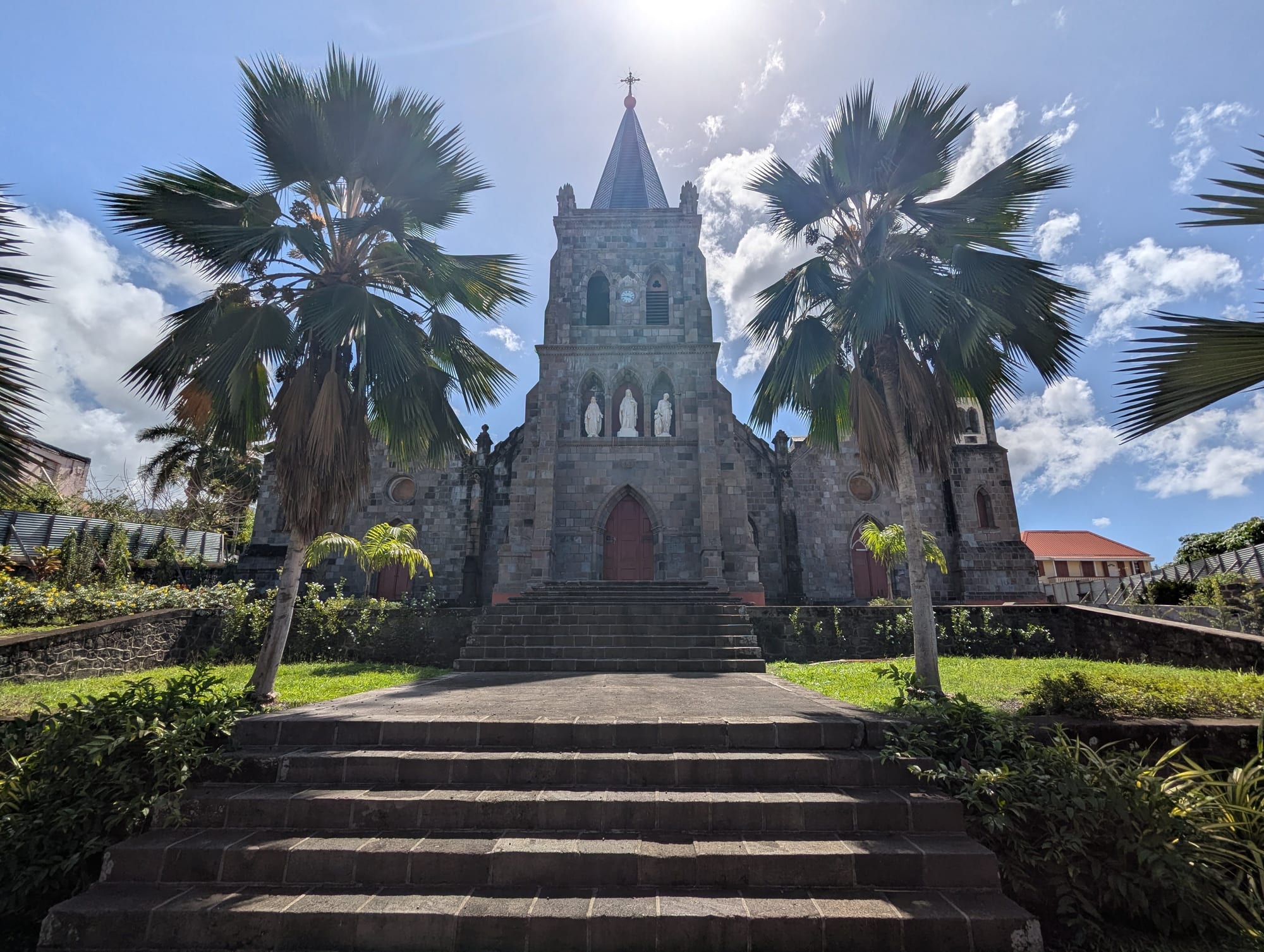 A grey stone Gothic-style cathedral with a central tower and spire and three white statues above the red door, reached by a long flight of steps between two tall palms