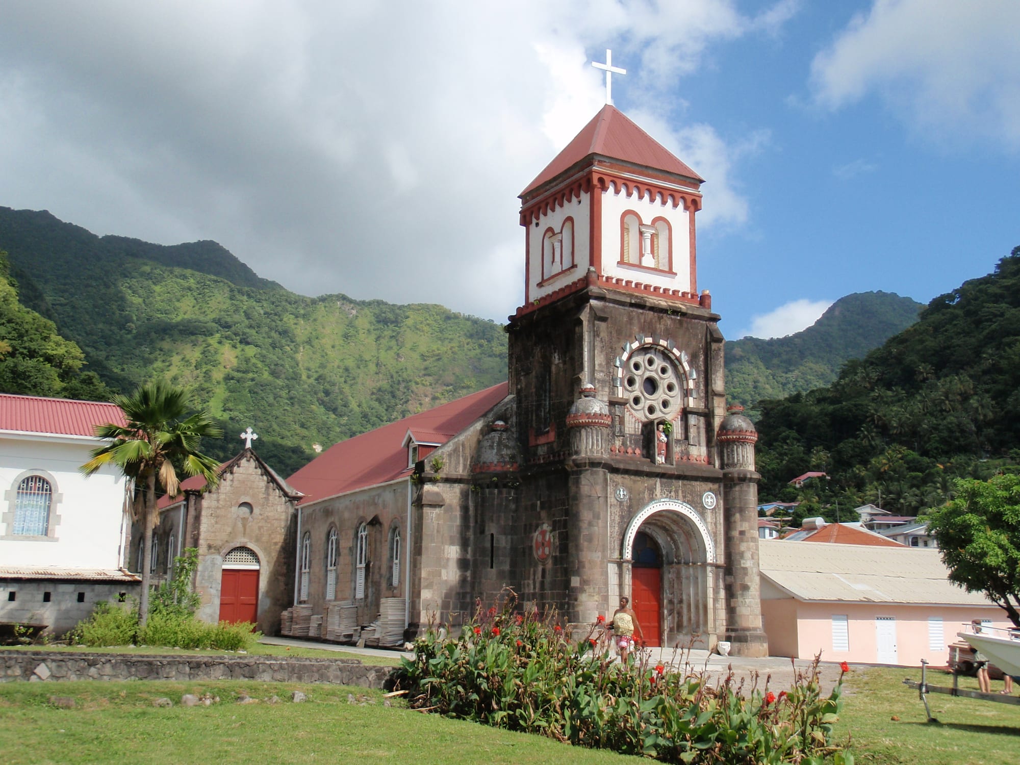 A dark stone church with a red-roofed bell tower topped by a cross, a rose window and a red arched door, set among red flowers and lawns below steep green mountains