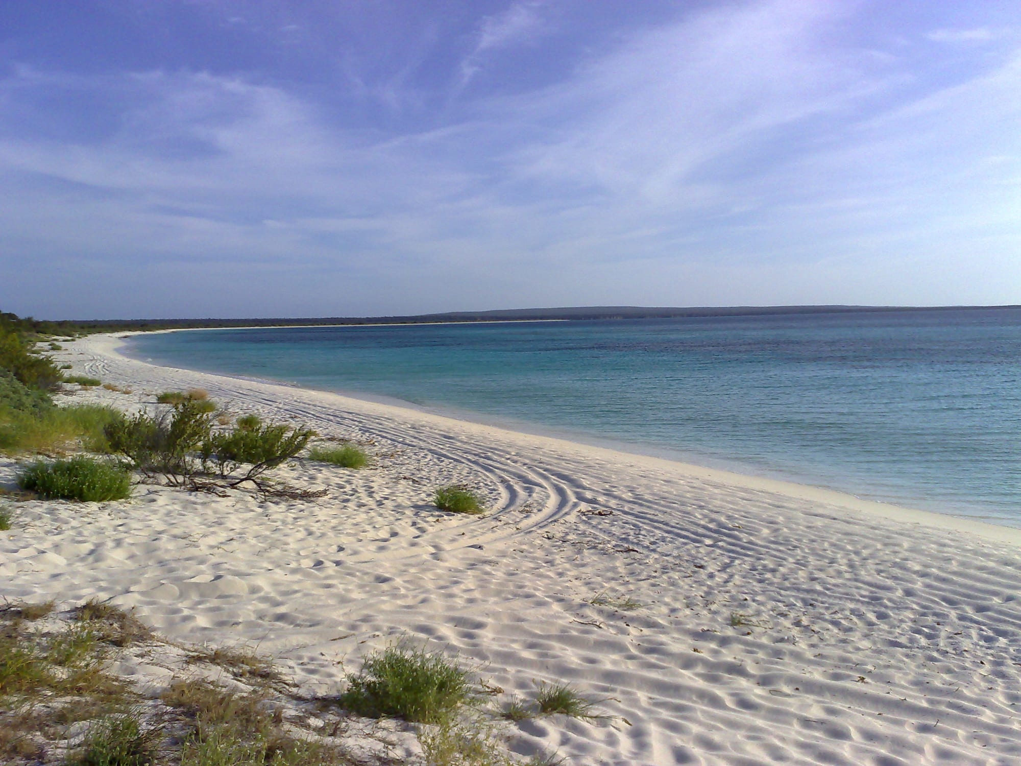 An arc of pale sand and shallow turquoise water below scrubby desert hills, completely empty