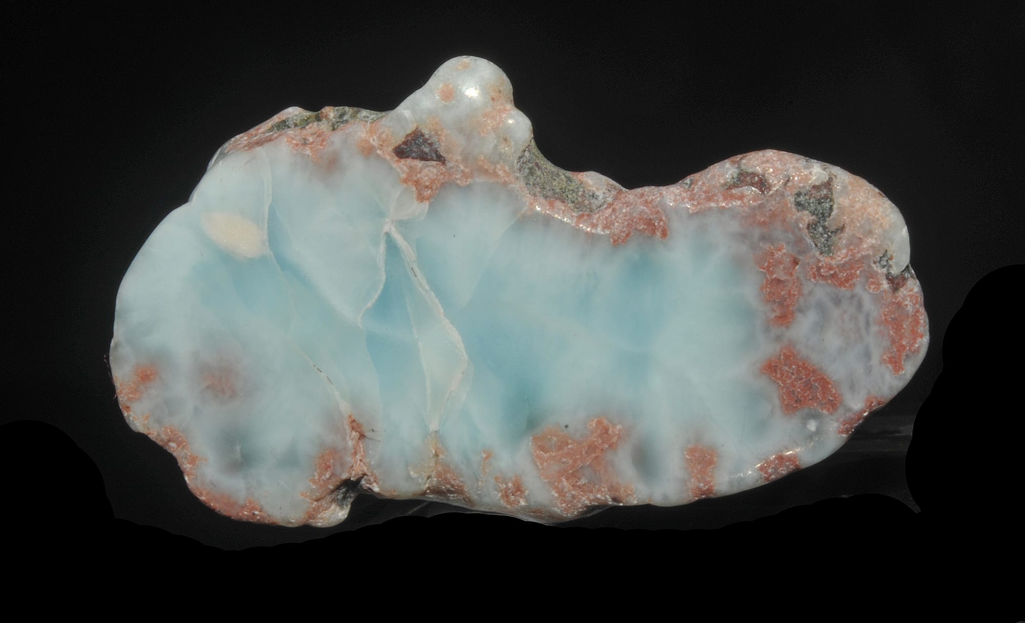 A polished, irregularly shaped piece of pale sky-blue stone with white veining and patches of reddish-brown rock around its edges, set against a black background