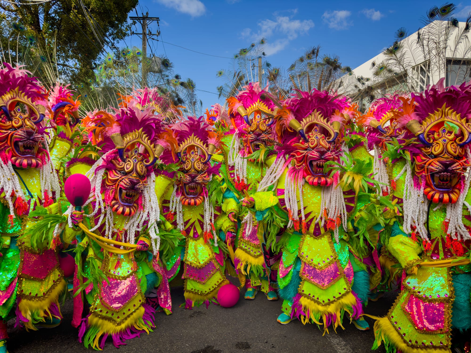 A tightly packed group of carnival performers in matching lime-green, pink and gold costumes with feathered headdresses and fanged, horned devil masks, standing in a street under a blue sky