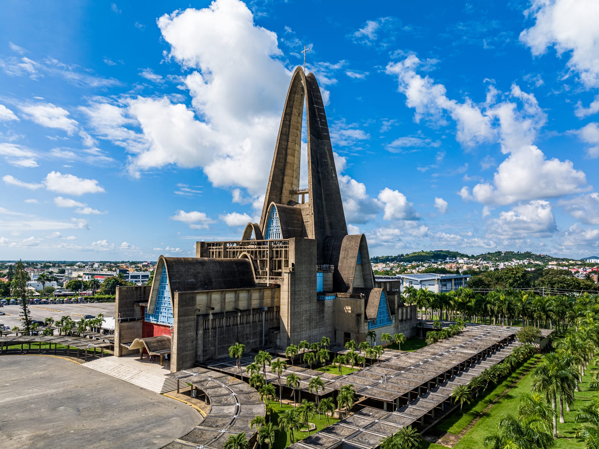 An aerial view of a modernist concrete basilica with a soaring pointed arch rising above lower arched wings, with blue glass panels, surrounded by palm-lined walkways and lawns