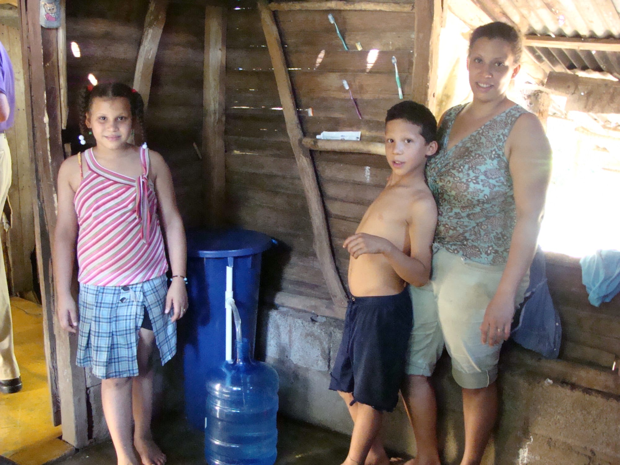 A mother and her two children standing beside a large water jug inside a wooden home in the Dominican countryside
