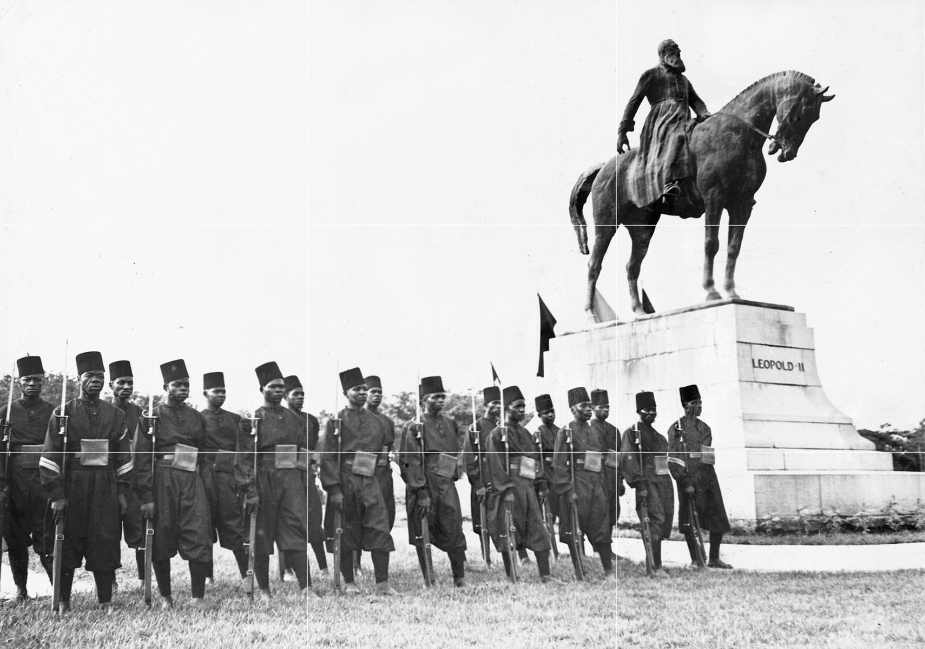 A colonial-era photograph of ranked African soldiers in fezzes standing before a statue of a European king