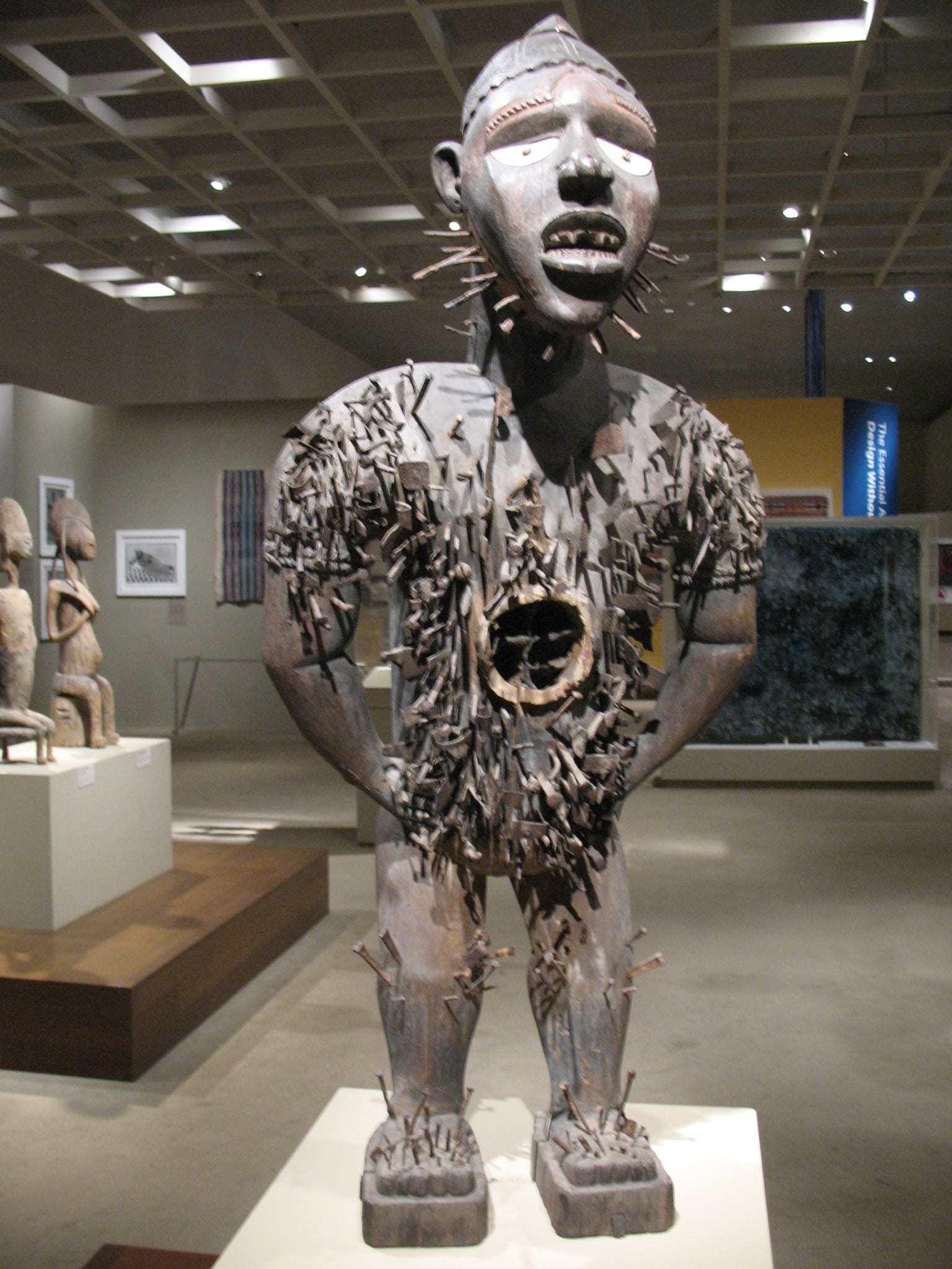 A carved wooden standing figure studded with hundreds of iron nails and blades, with a fierce expression