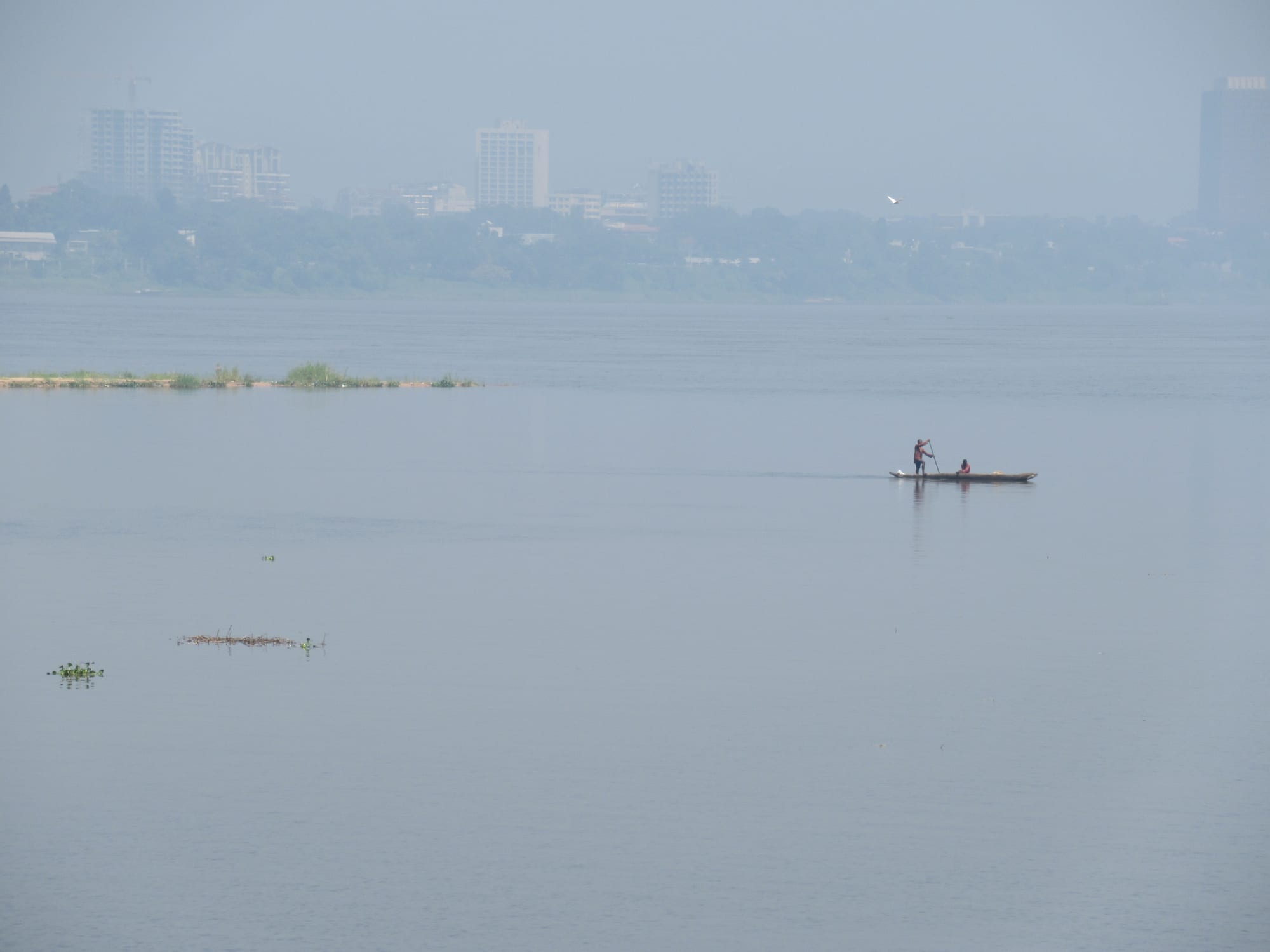 Two men poling a long wooden dugout canoe on a broad brown river bordered by forest
