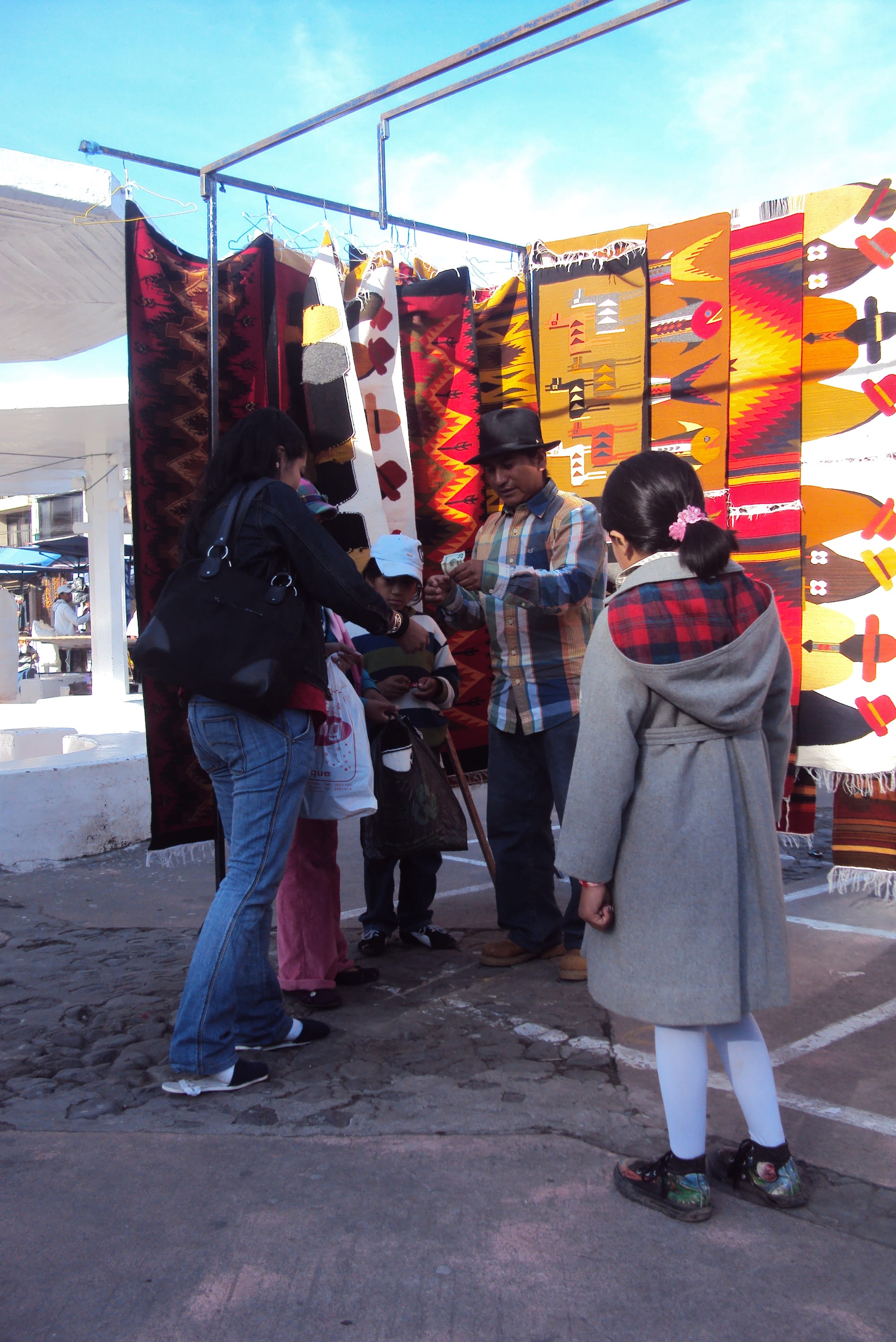 A family buying a hand-woven textile from a vendor at an outdoor market stall hung with colourful patterned rugs