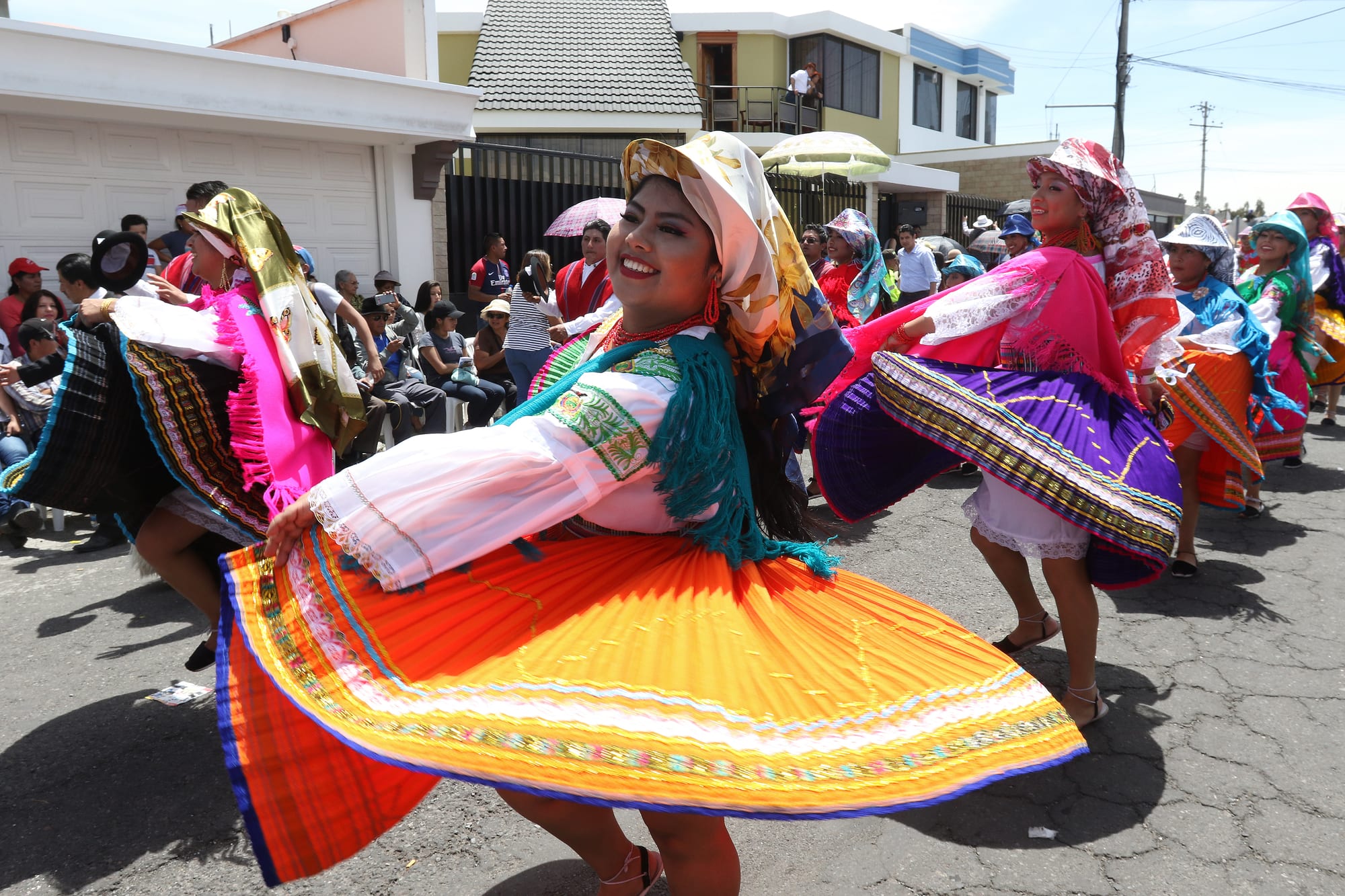 A smiling woman in a white embroidered blouse, a fringed shawl and a headscarf swirls a wide orange pleated skirt as she dances in a street, with other dancers in bright skirts and spectators behind her