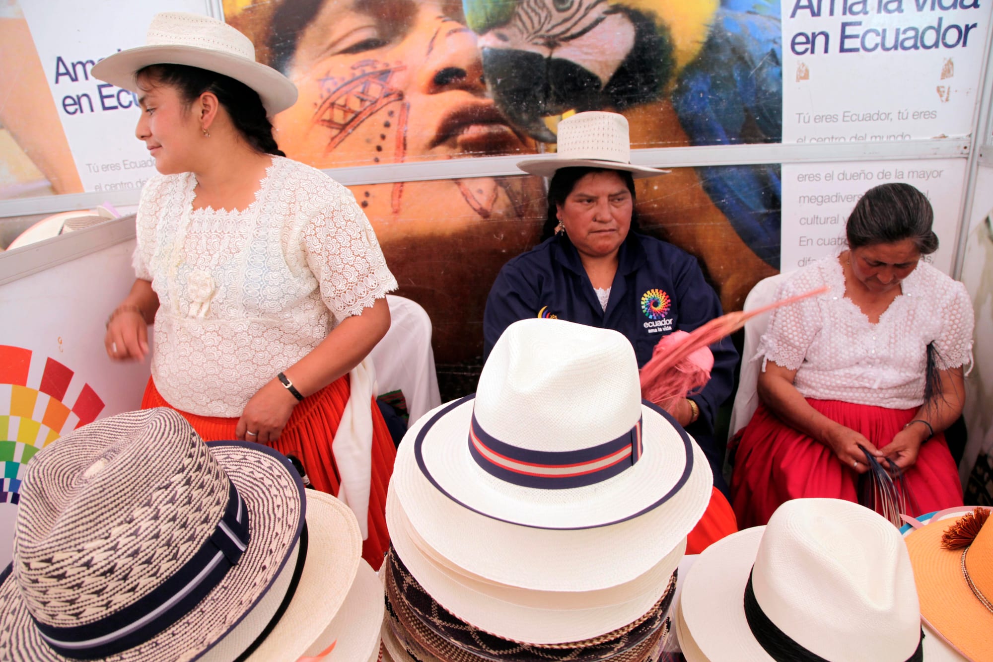 Three women at a craft stall behind stacks of white and patterned straw hats; the woman on the right is weaving strands of straw in her lap