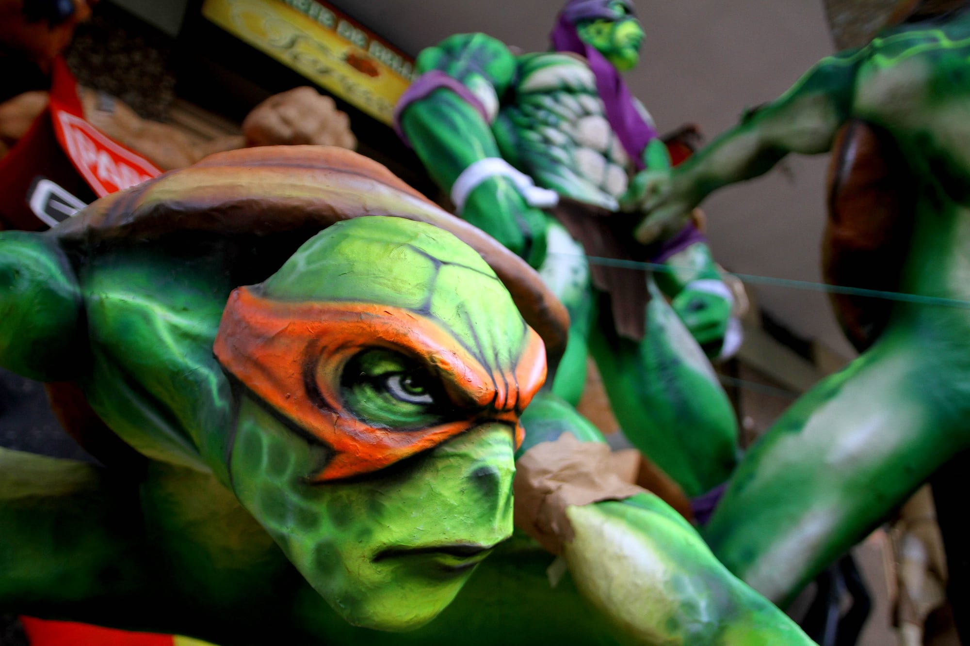 A close-up of a large painted papier-mâché effigy of a green cartoon turtle with an orange eye mask, with other brightly painted effigies behind it