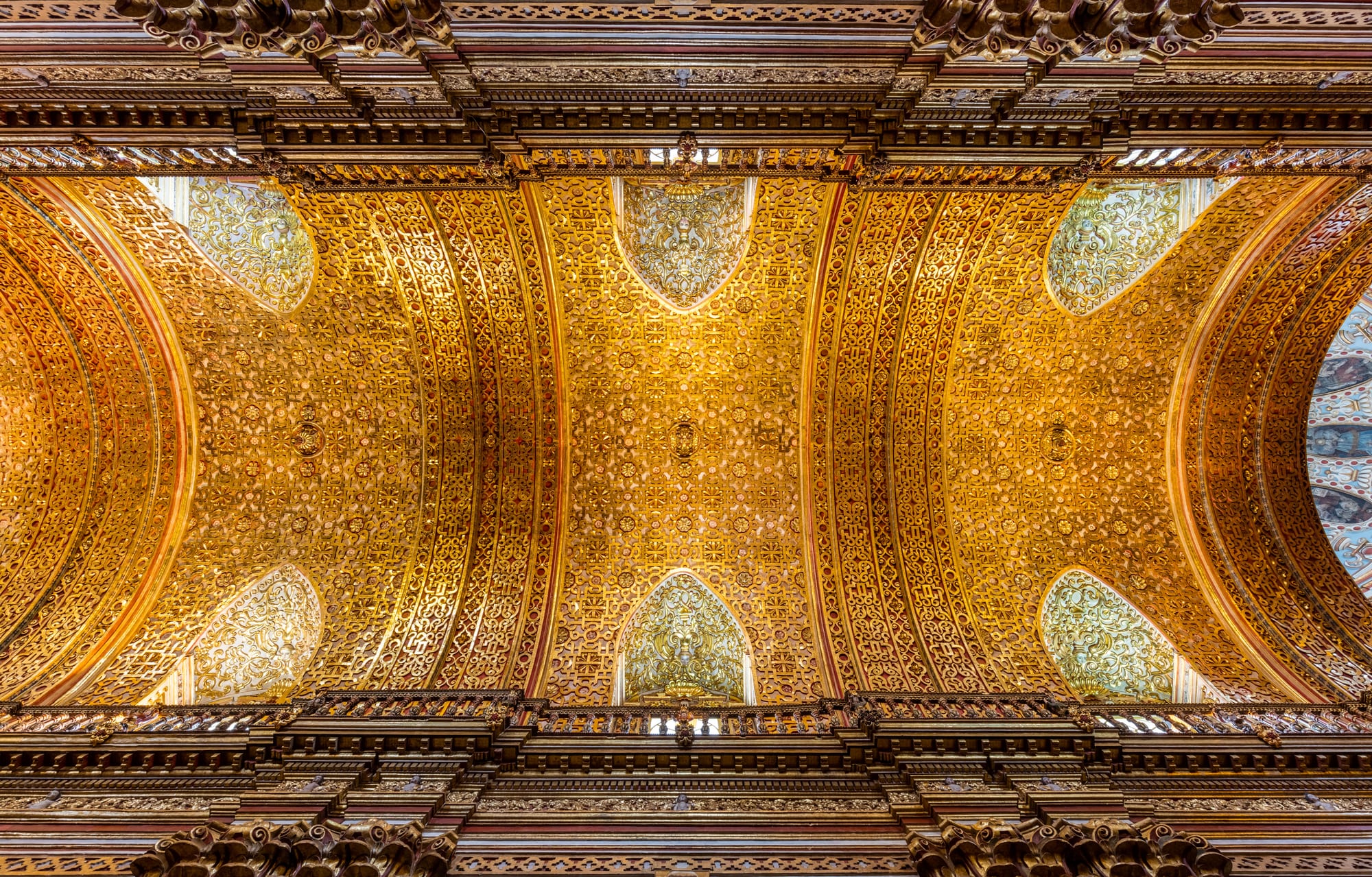 Looking straight up at a barrel-vaulted church ceiling covered in intricate gilded geometric patterns, framed by carved wooden cornices and pale arched windows