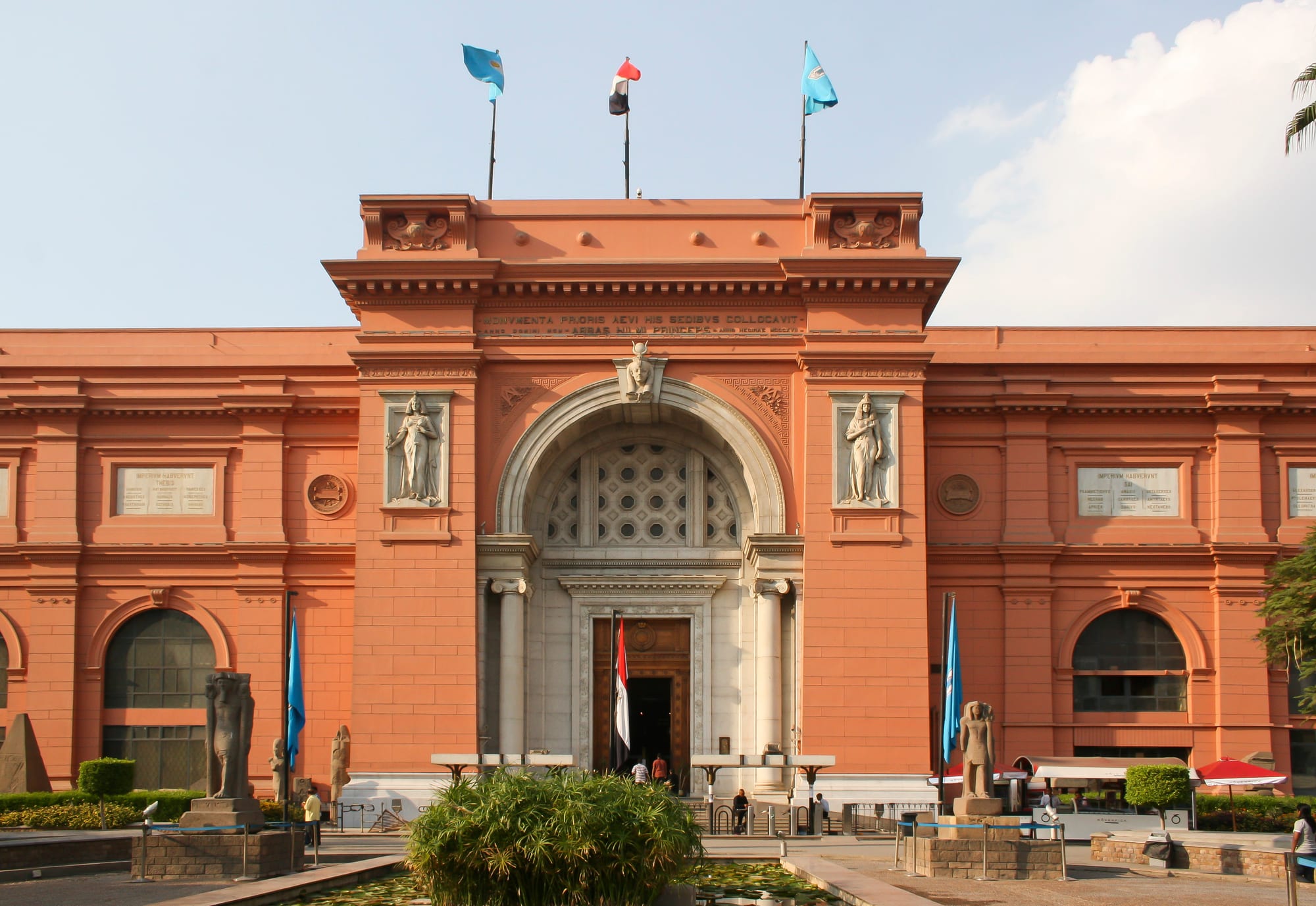 A grand pink neoclassical museum building behind a garden and palm trees