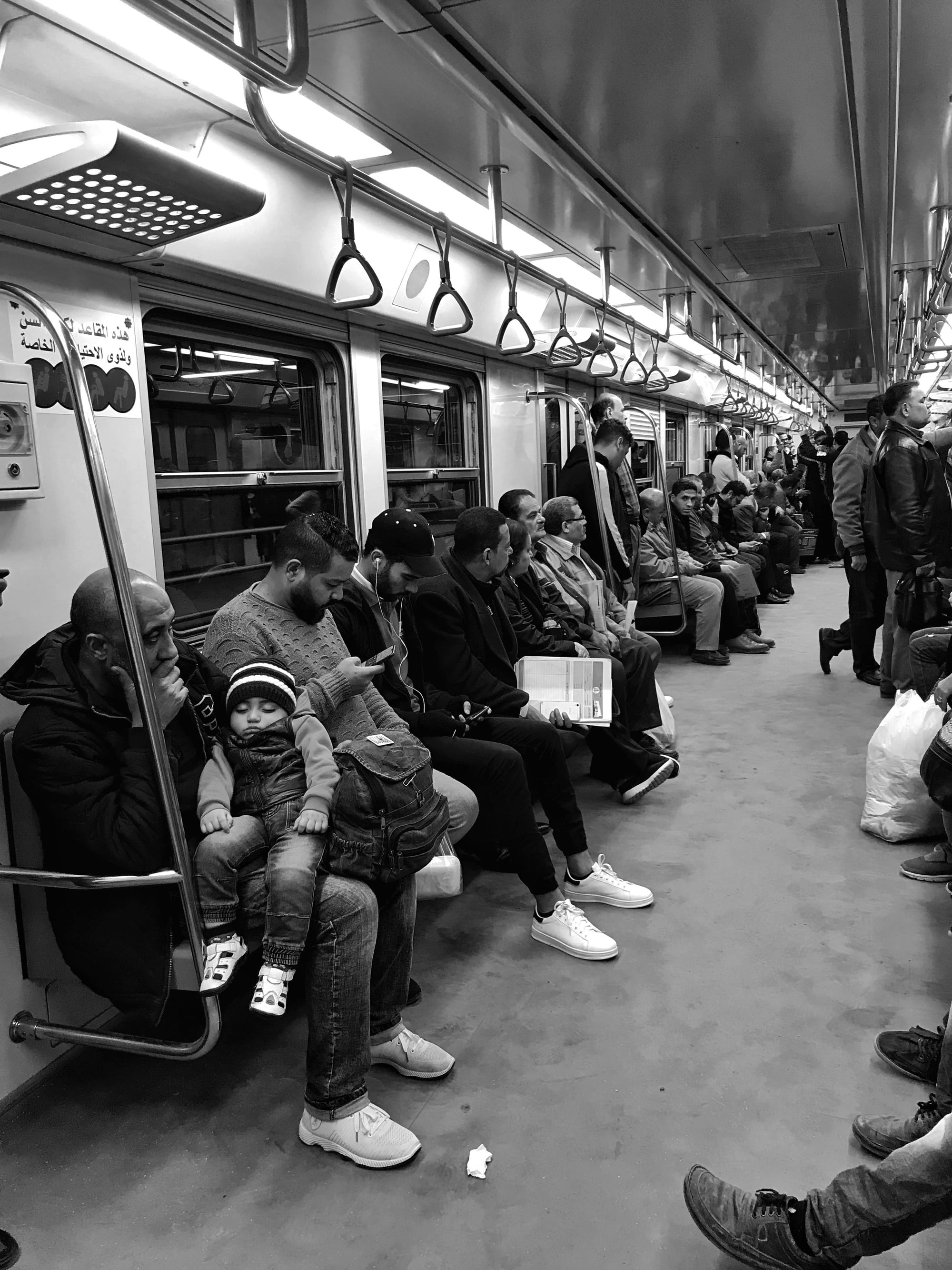 Black and white photograph of a crowded Cairo metro carriage, a man holding a sleeping toddler among seated and standing passengers